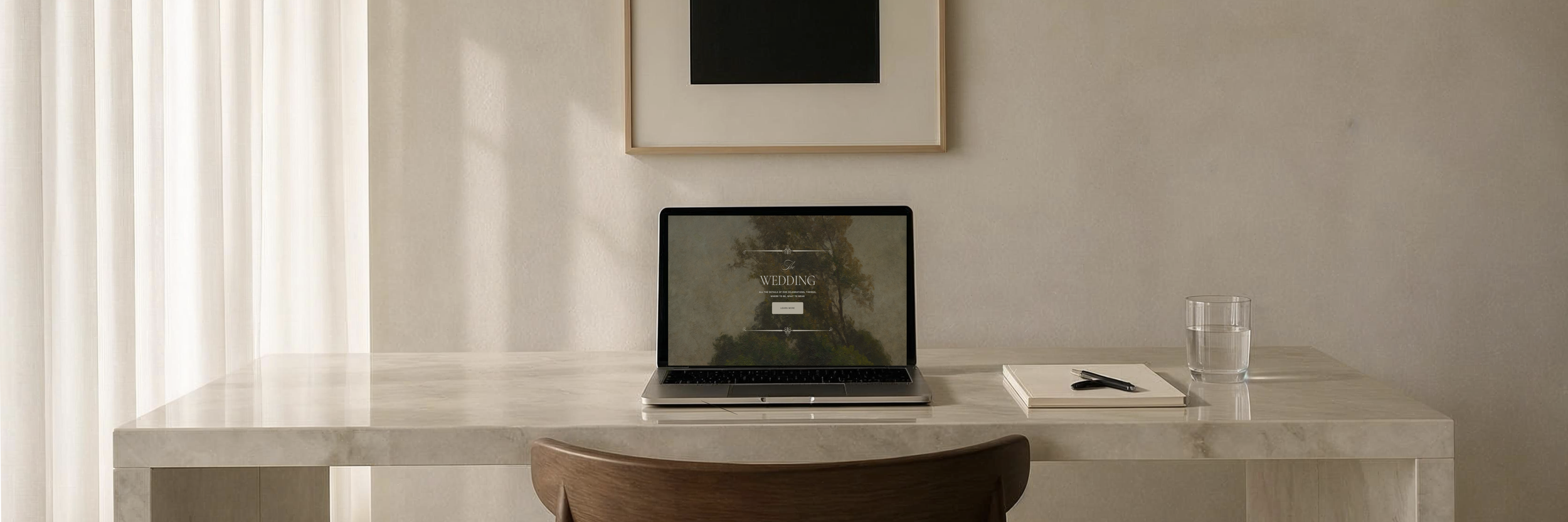 A minimalist desk with a laptop displaying a wedding website, a glass of water, a notebook, and a pen, with a neutral-toned wall and cream-colored curtains in the background.