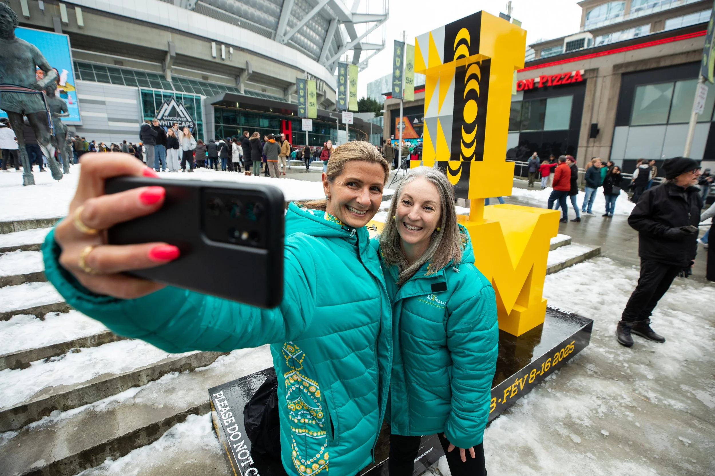 Two smiling women in blue jackets taking a selfie in front of a yellow Vancouver 2010 Olympics promotional sculpture outside a stadium, with snow on the ground and a crowd in the background.