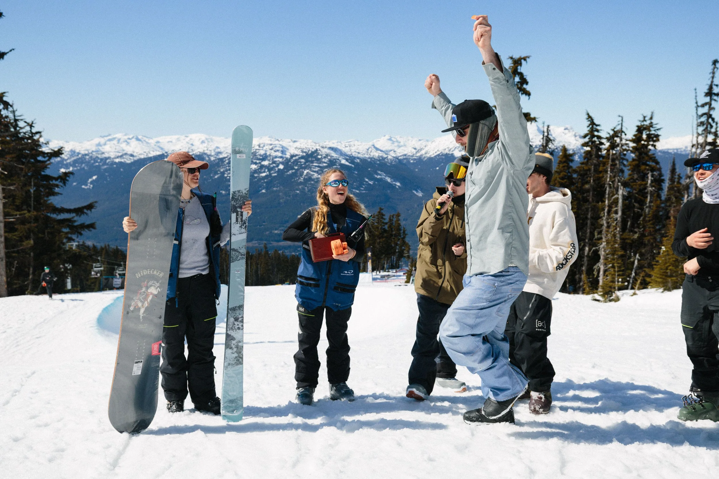 Group of people in winter clothing on snow, with mountains and pine trees in the background, some wearing goggles and hats, and a person holding a snowboard and snowboard poles.
