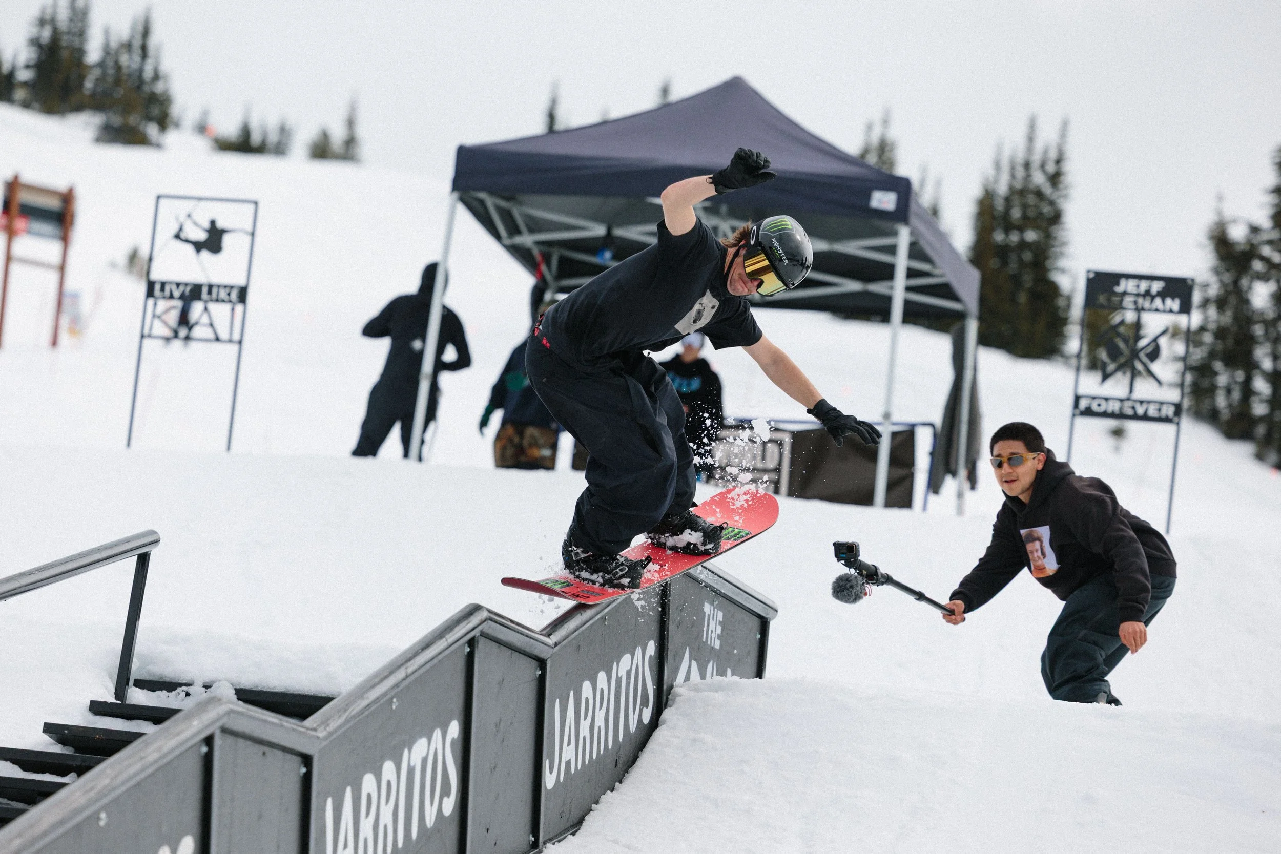 A snowboarder wearing a black helmet and black clothing is mid-air performing a trick on a rail at a snowy ski resort. There is a tent and skiers in the background, along with trees and signage that includes the words "LIVE LIKE" and "JARI POTS".