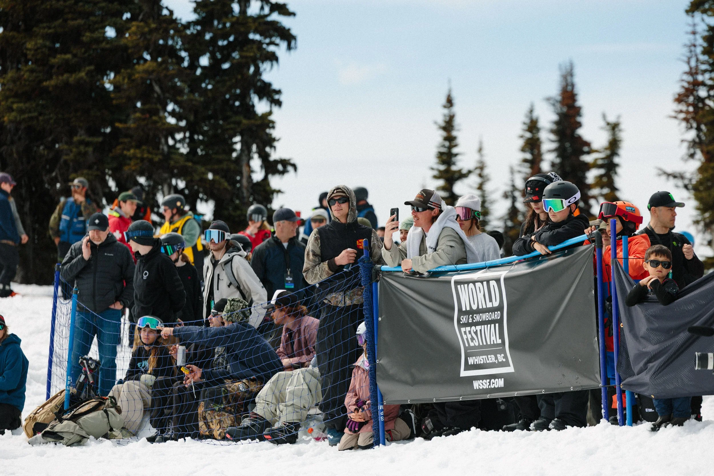 A group of people on snow gear gathered at a ski festival, some standing and some sitting behind a blue safety fence with a banner reading 'World Ski & Snowboard Festival' in Whistler, BC, surrounded by snow-covered ground and tall pine trees under a