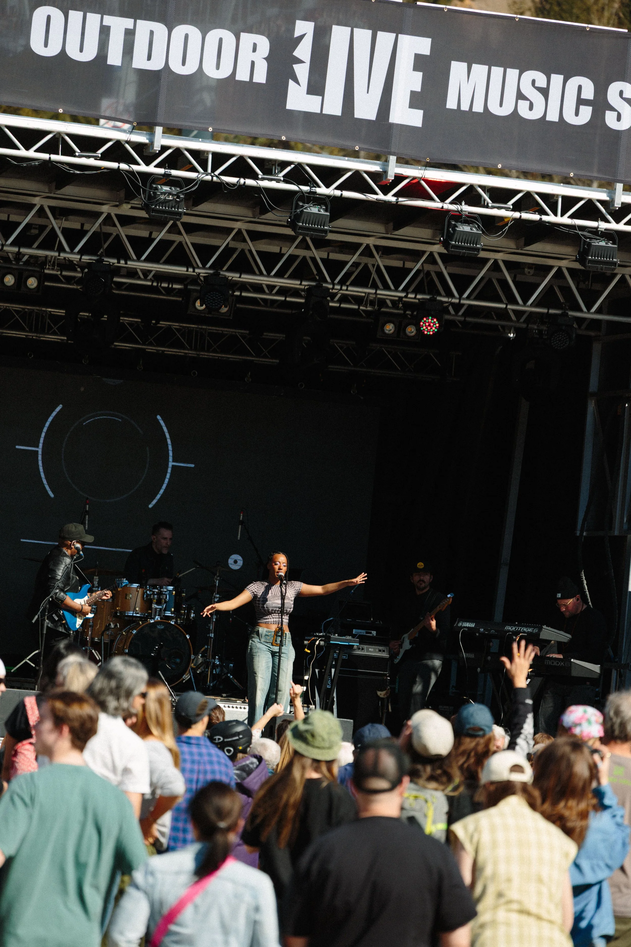 A live outdoor music performance with a female singer, a band on stage, and a crowd of audience members watching.