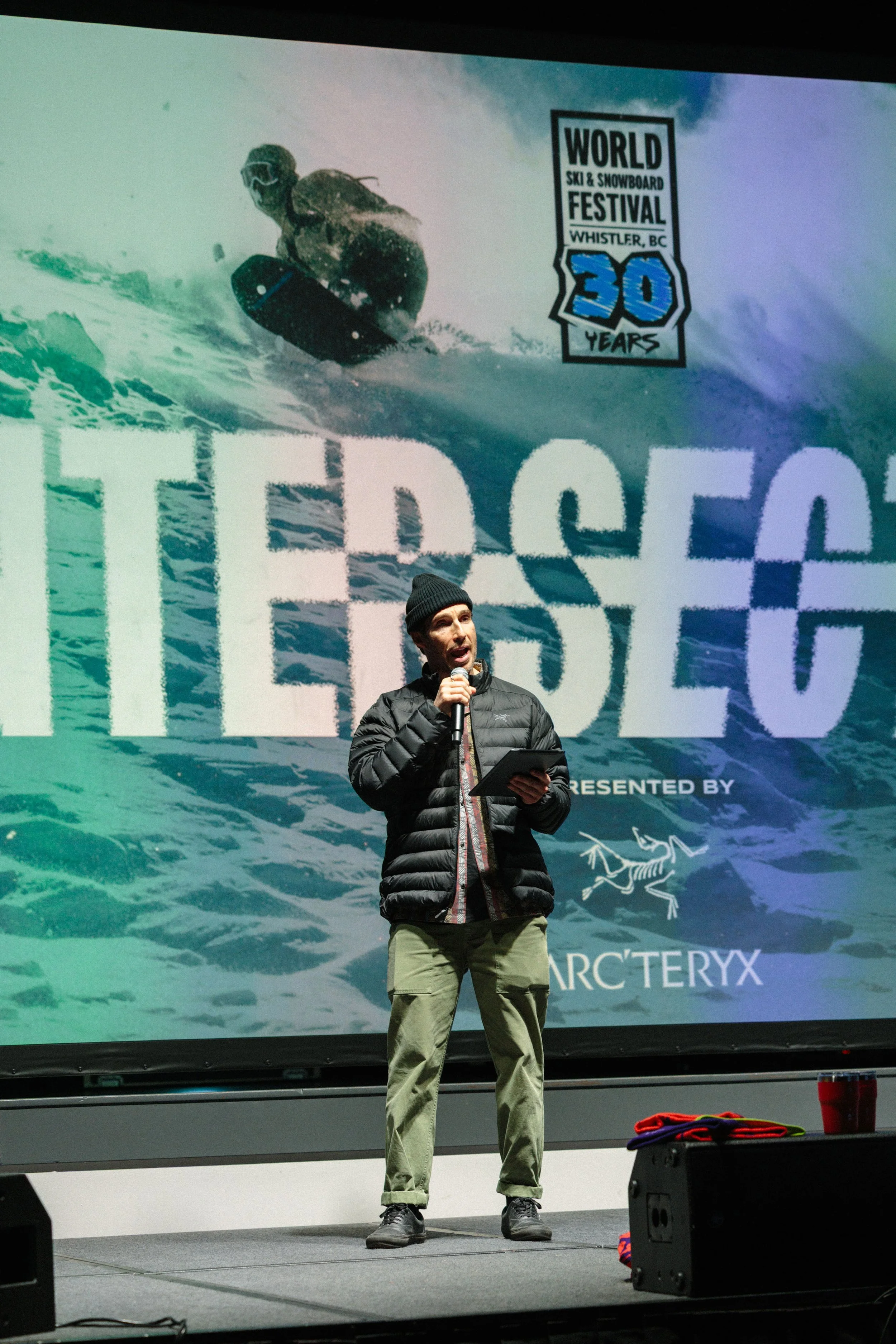 A man in a black jacket, green pants, and a black beanie speaking into a microphone on a stage, with a large screen behind him showing an image of a snowboarder and the text 'World Ski & Snowboard Festival Whistler, BC 30 Years' and 'Archery' with a 