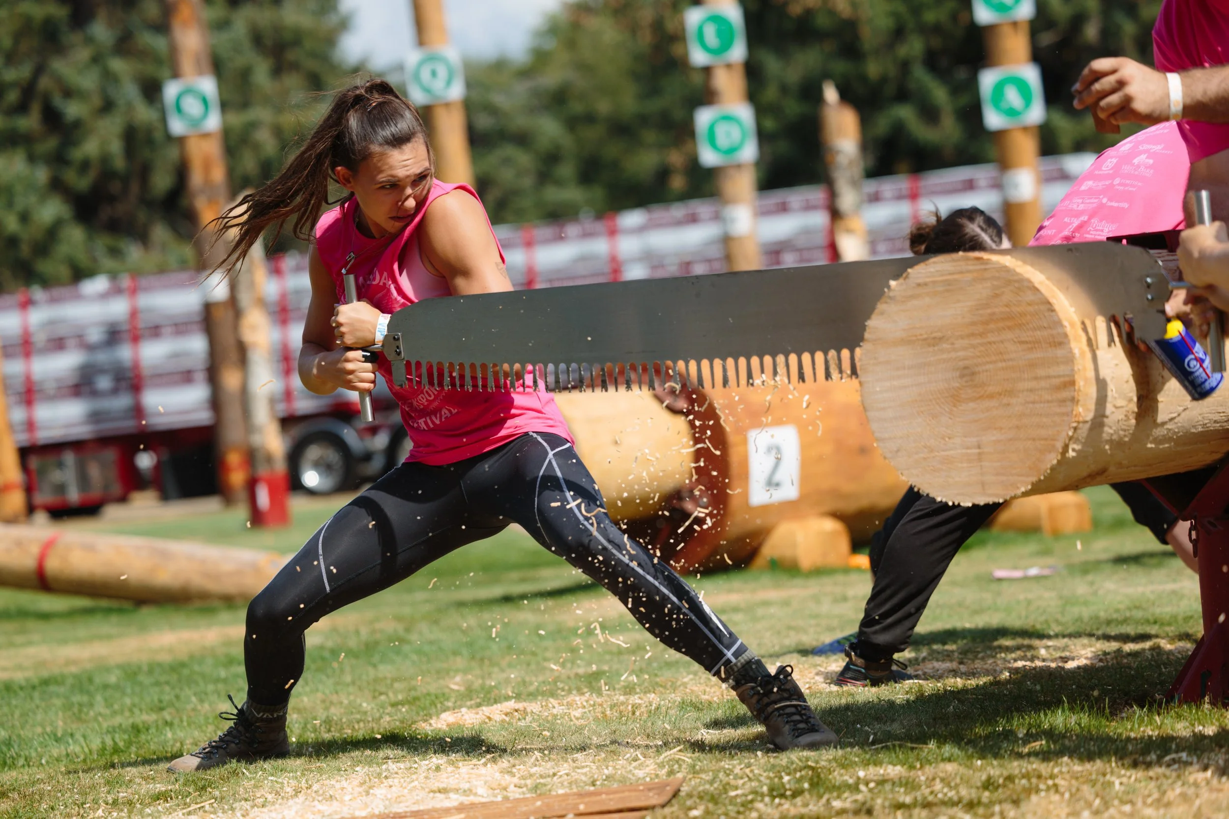 Female competitor in a pink tank top and black leggings uses a large saw to cut through a log during a lumberjack competition, with sawdust flying and onlookers nearby.