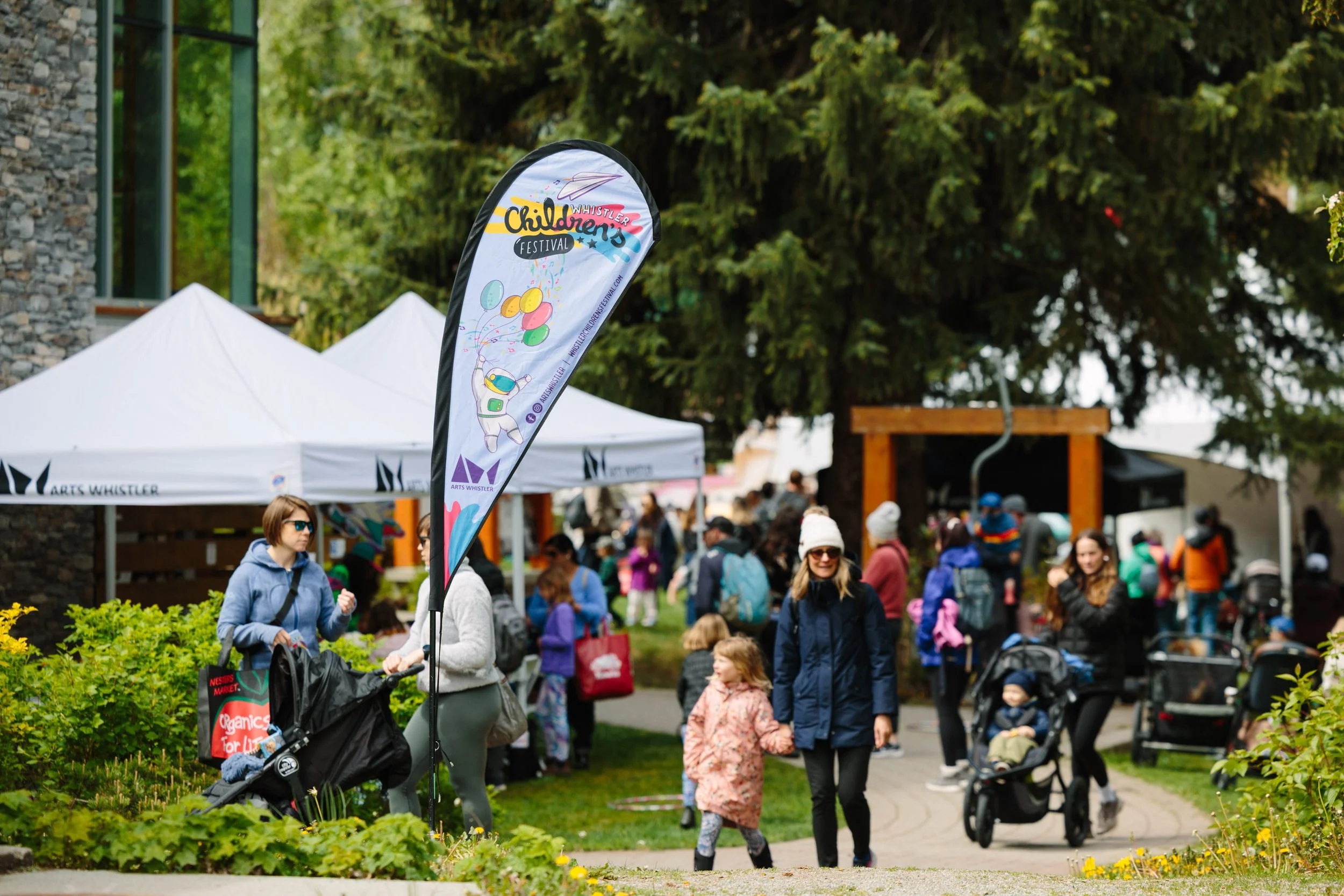 People walking and gathering at an outdoor festival with tents and banners, including a flag titled 'Whistler Children’s Festival'. Children and adults are wearing jackets and hats, some pushing strollers. The setting is lush with trees and greenery.