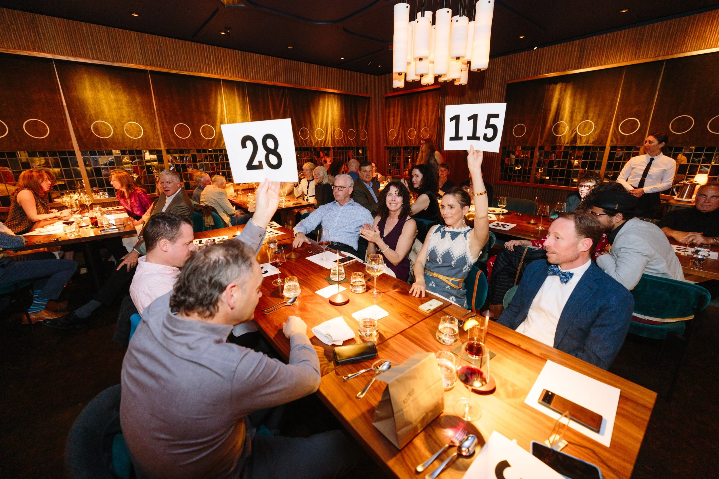 People attending an auction at a restaurant, with two women holding up numbered paddles, 28 and 115, surrounded by glasses of wine and dinnerware on the table.