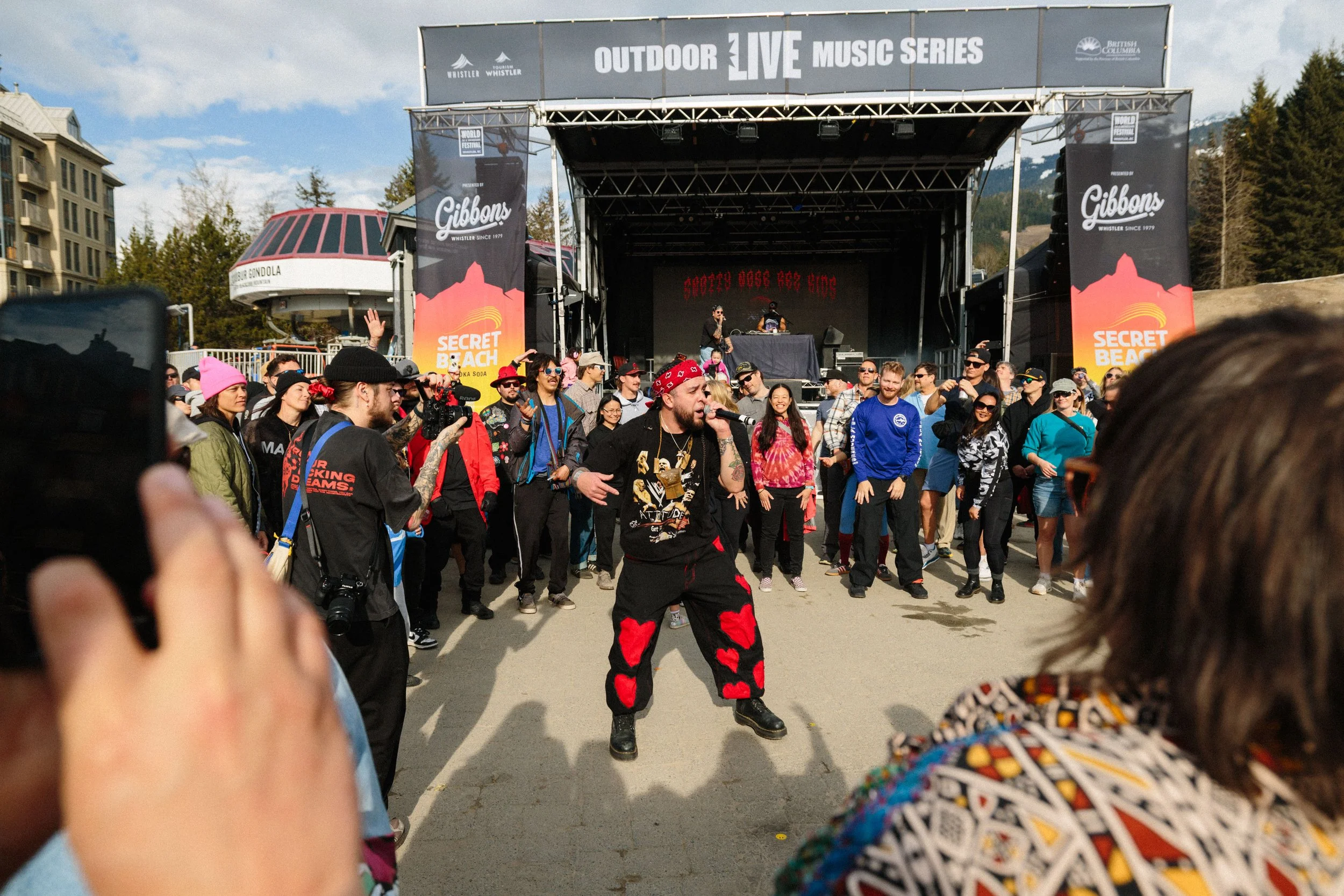 Crowd of people gathered in front of an outdoor stage at a music festival, with a DJ and singer performing. The stage has banners advertising 'Outdoor Live Music Series' and 'Secret Beach,' with a backdrop of trees and mountains.