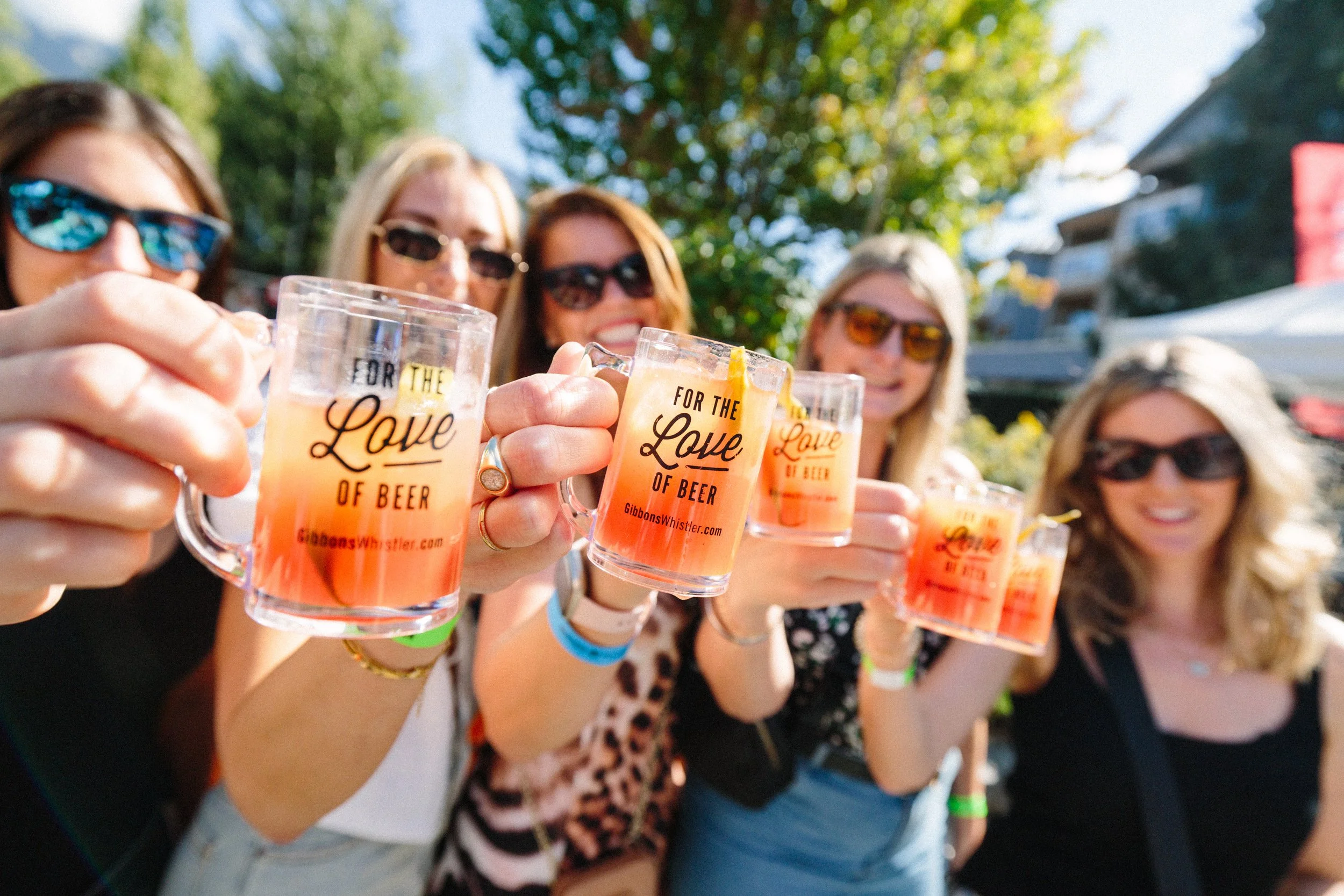 Group of six women outdoors, holding glasses with a pink drink, celebrating and smiling at the camera.