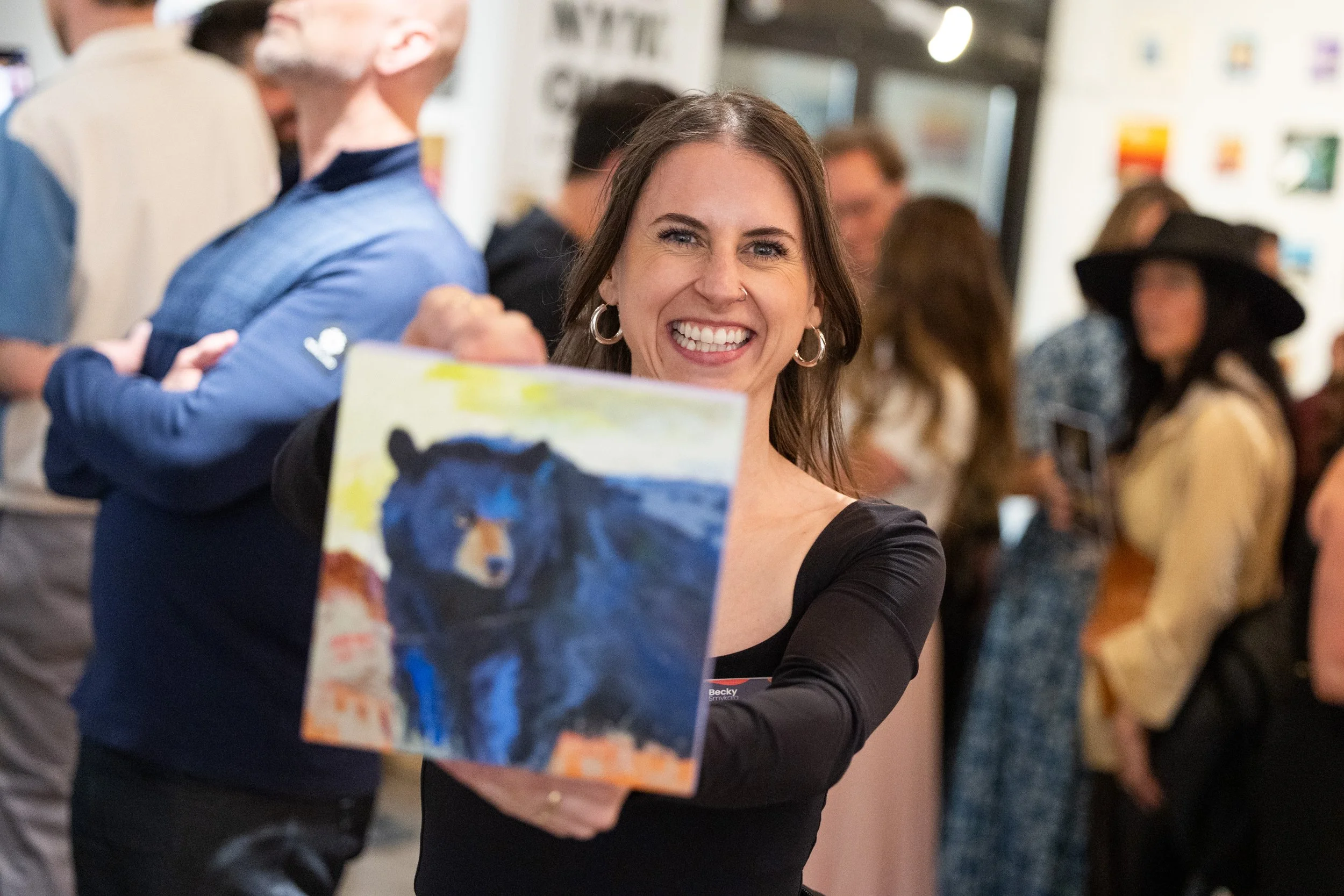 A woman smiling and showing a painting of a black bear with a mountainous landscape in the background at an art gallery or exhibition.