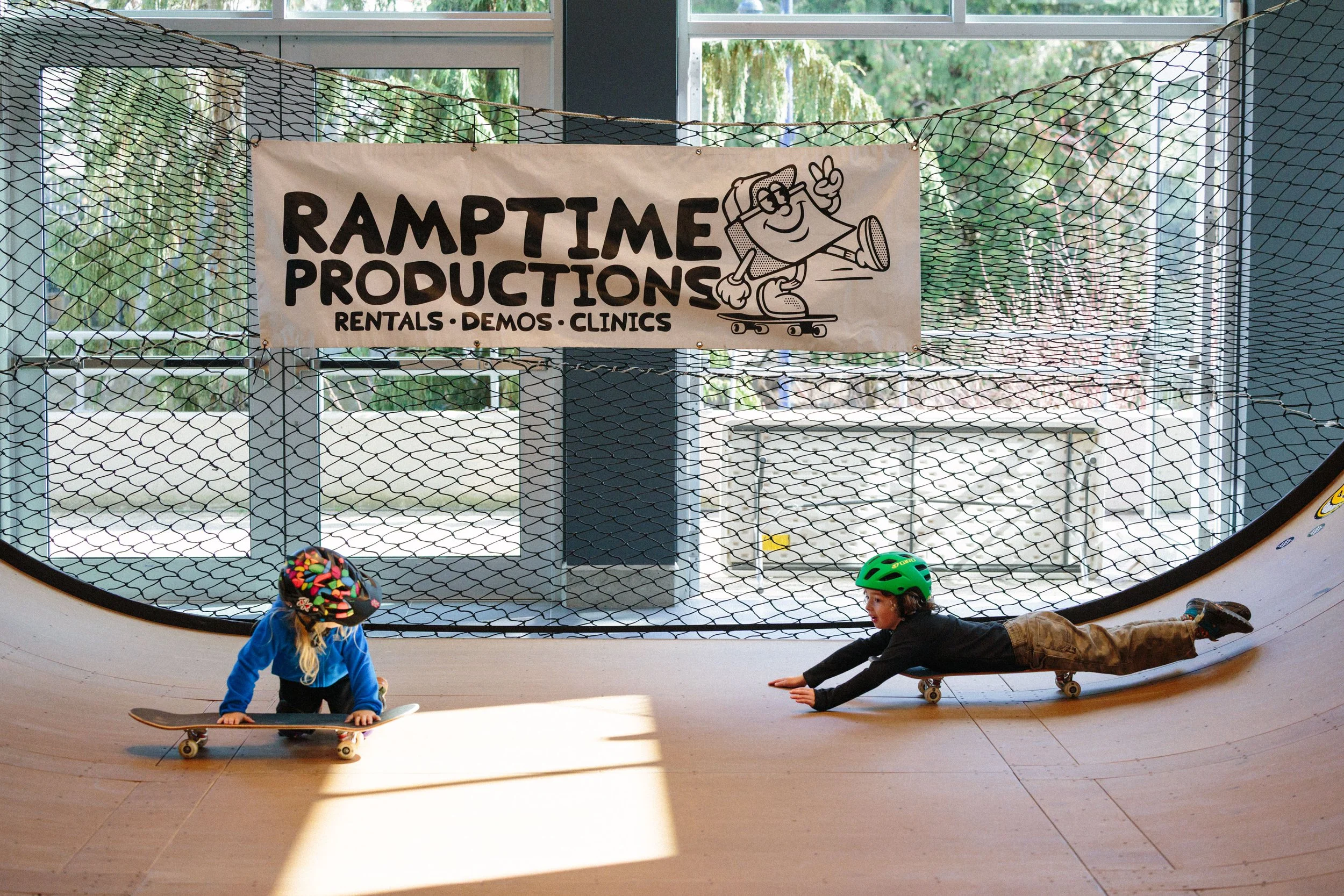 Children skateboarding inside a skate park with a sign that reads "Ramtime Productions Rentals Demos Clinics" and a cartoon character skateboarding.