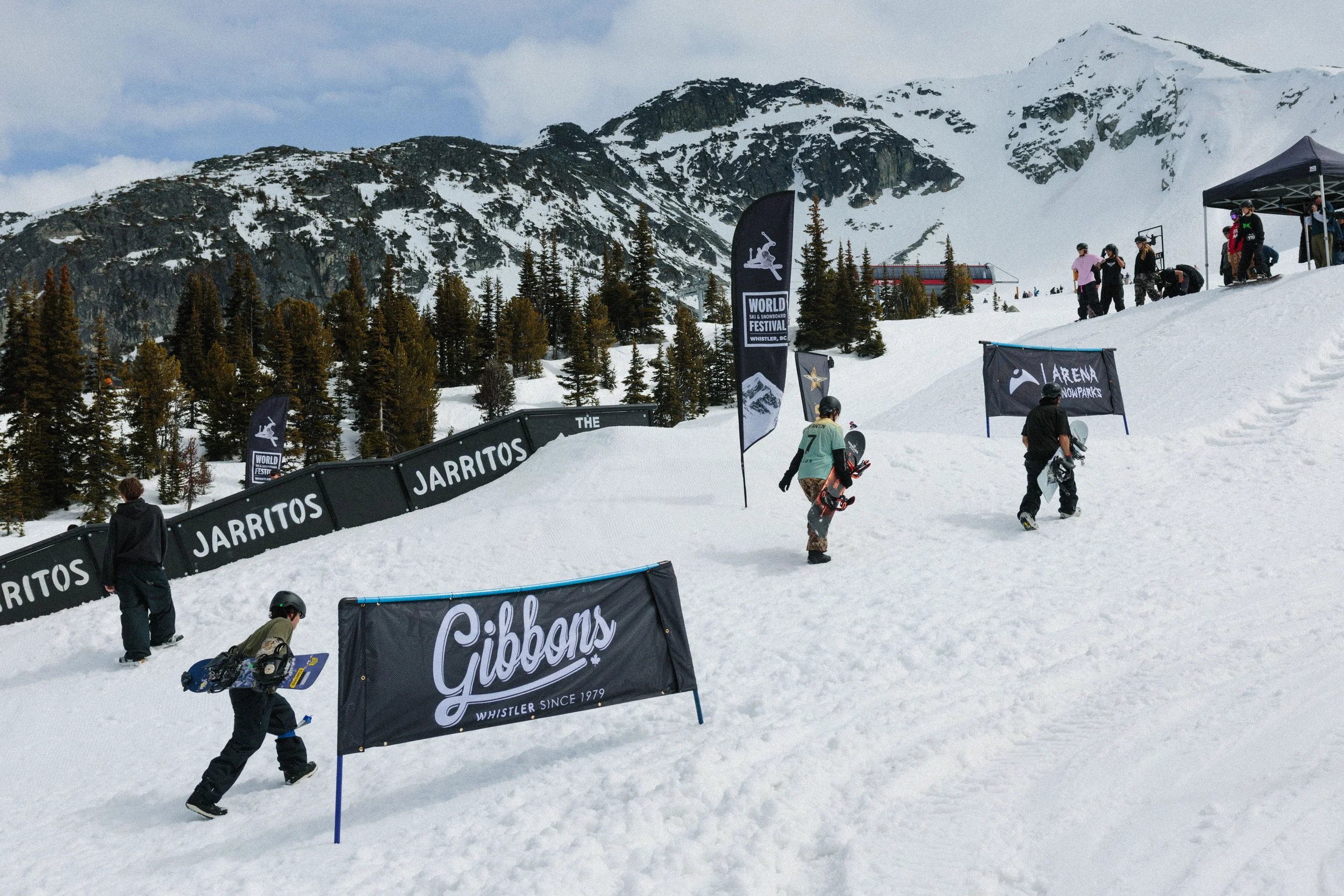 People skiing and snowboarding on a snowy mountain slope at a ski festival in Whistler, British Columbia. There are flags and banners with logos, and a mountain with snow in the background.