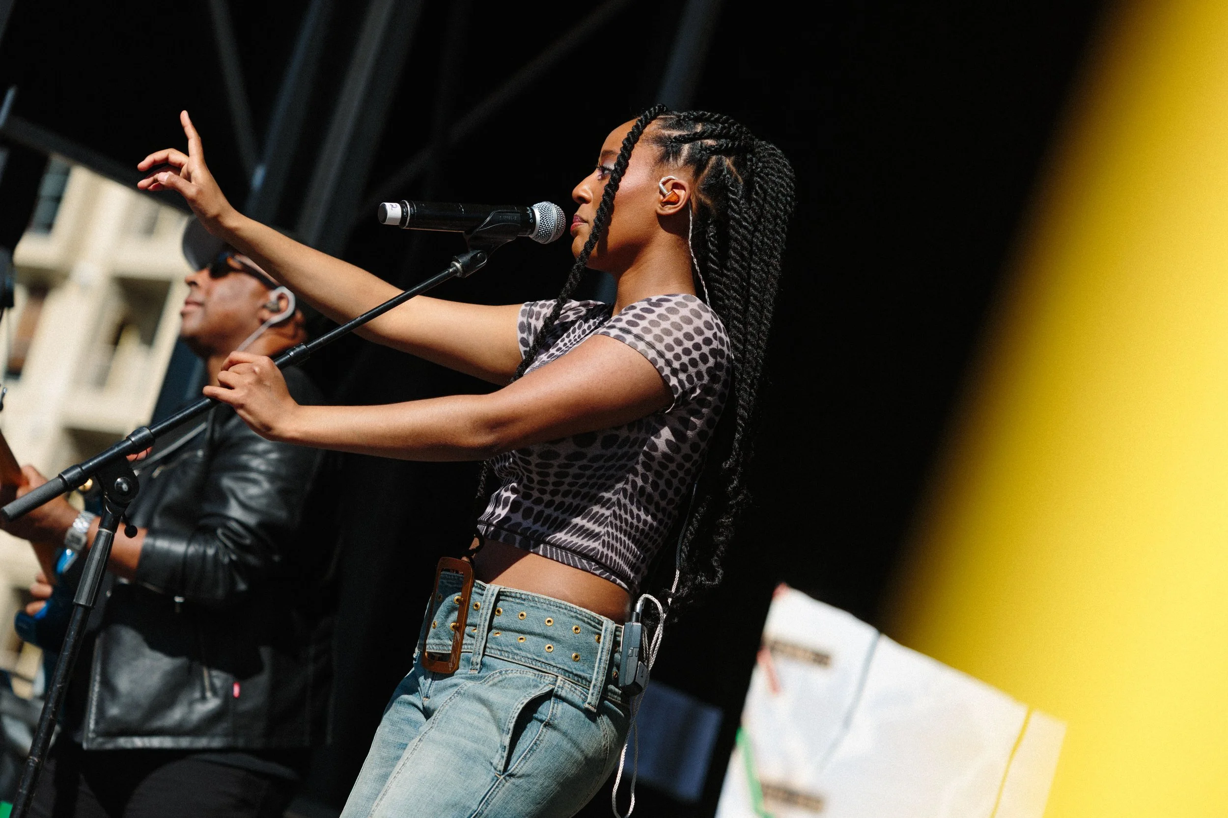 A woman with braided hair wearing a polka dot crop top and jeans vertical on stage speaking into a microphone, with a man in the background and a yellow object on the right side.