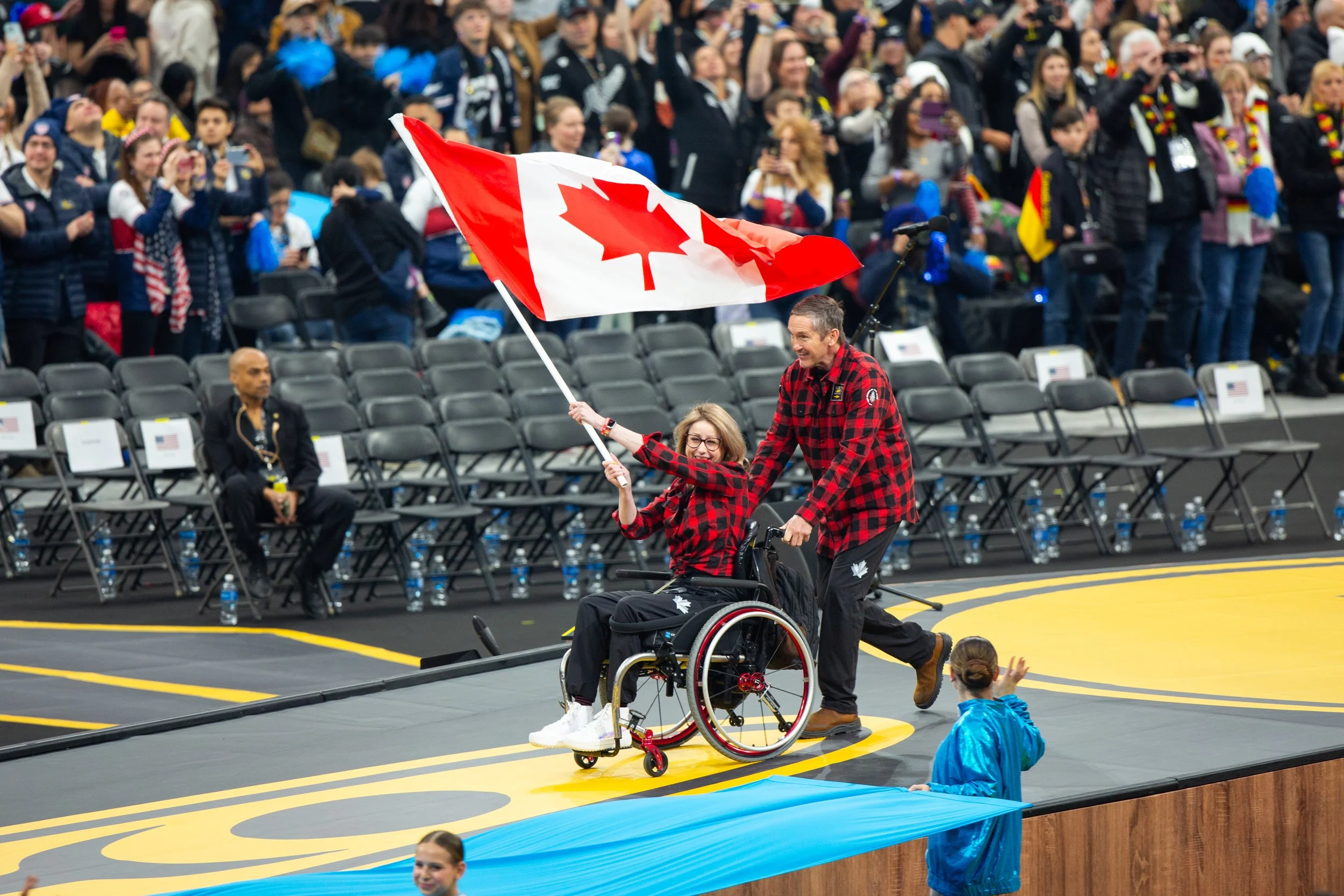 A woman in a wheelchair holding a Canadian flag, with a man assisting her, both wearing matching red and black plaid shirts, at an indoor sports event with a crowd in the background.