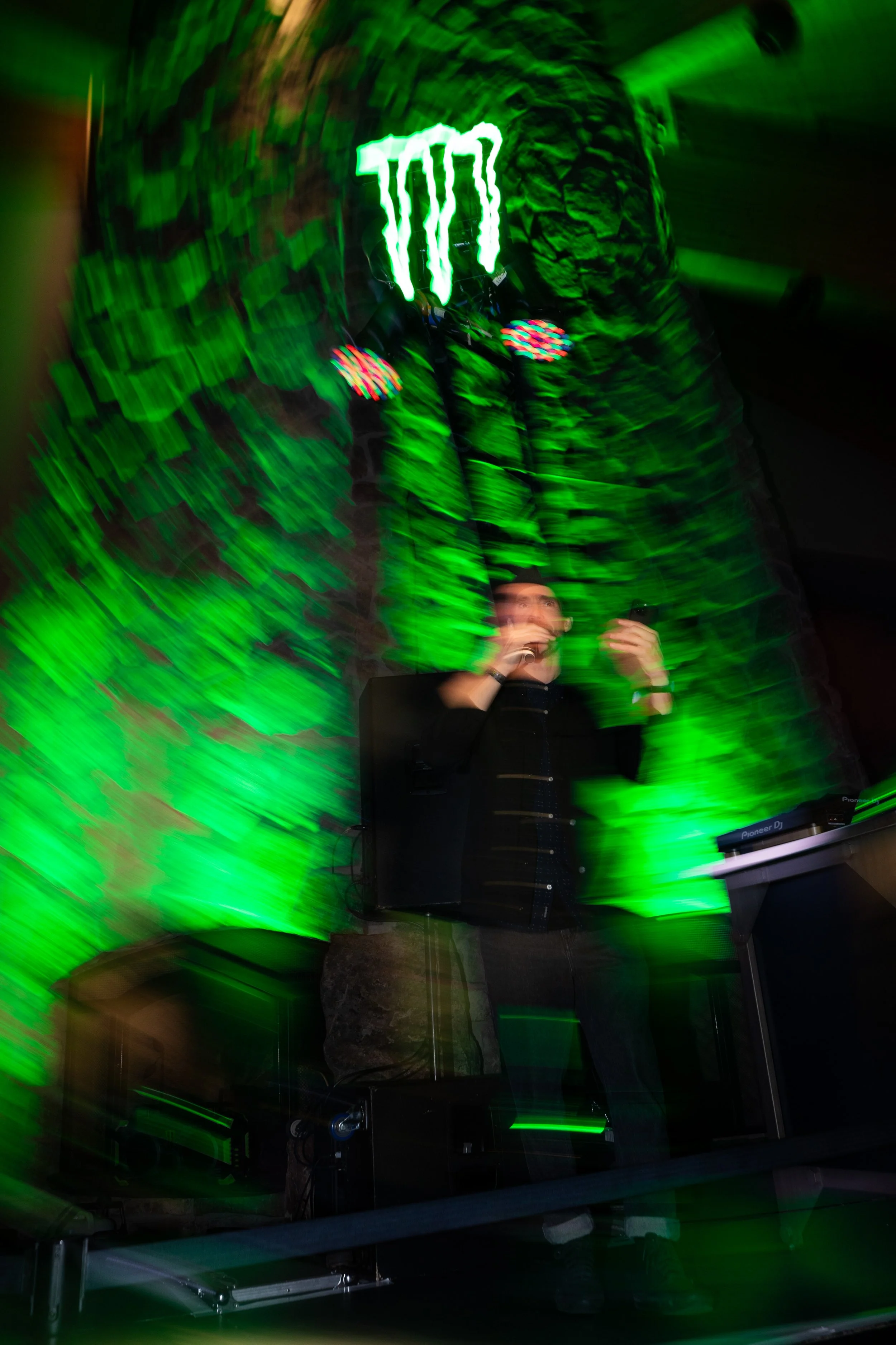 A person speaking or singing into a microphone at a nightclub or event with green lighting and a blurred motion effect.