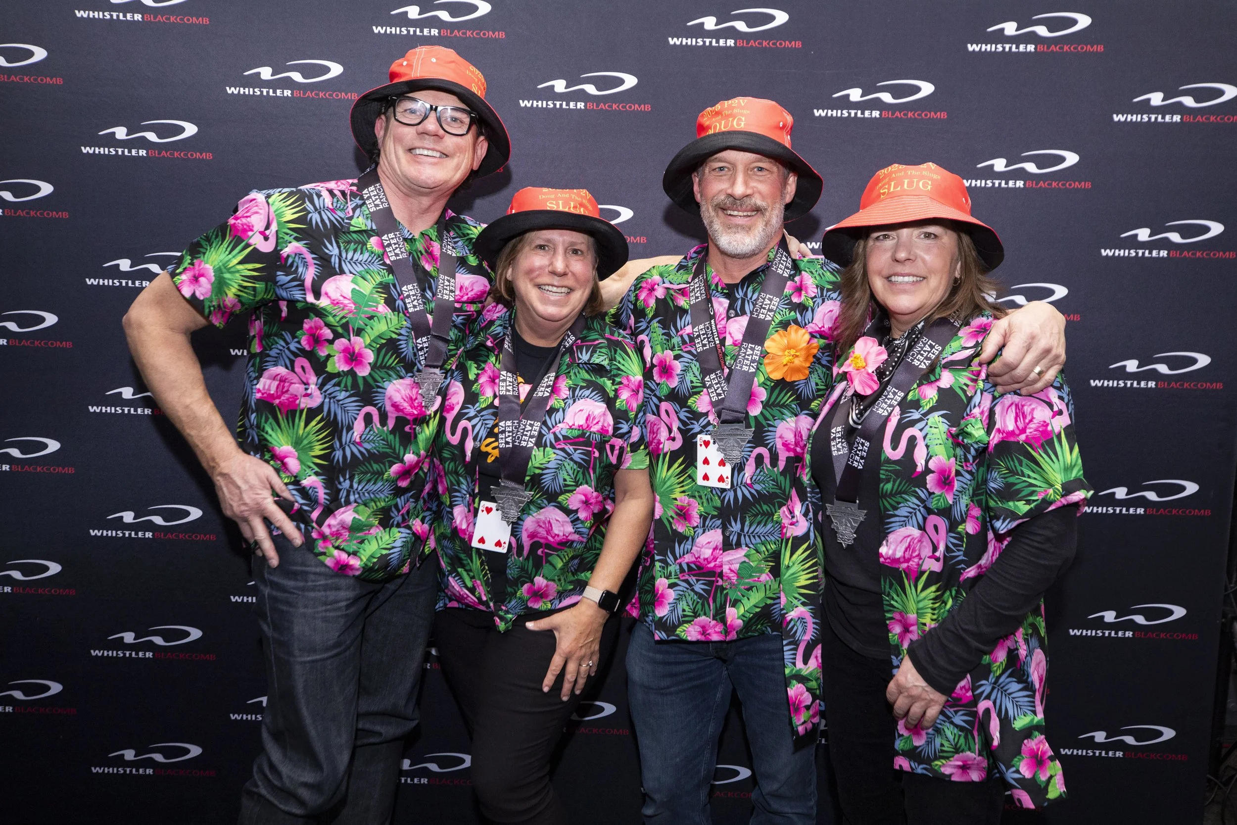 Four people at the Whistler Blackcomb event wearing matching Hawaiian shirts, bucket hats with 'SLUG' written on them, and medals around their necks, standing in front of a backdrop with the Whistler Blackcomb logo.