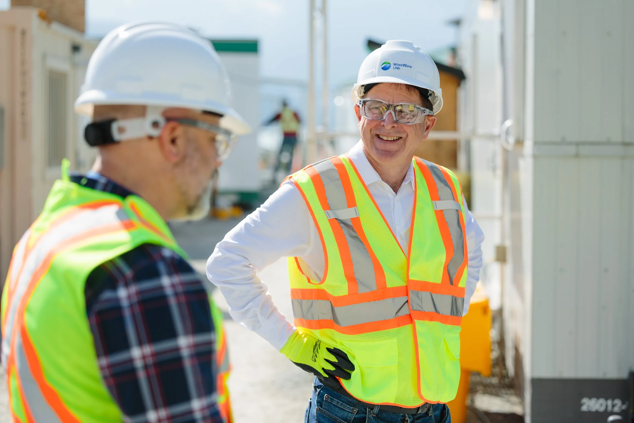 Two construction workers wearing safety helmets, high-visibility vests, and safety glasses, standing outdoors at a construction site and engaging in conversation.