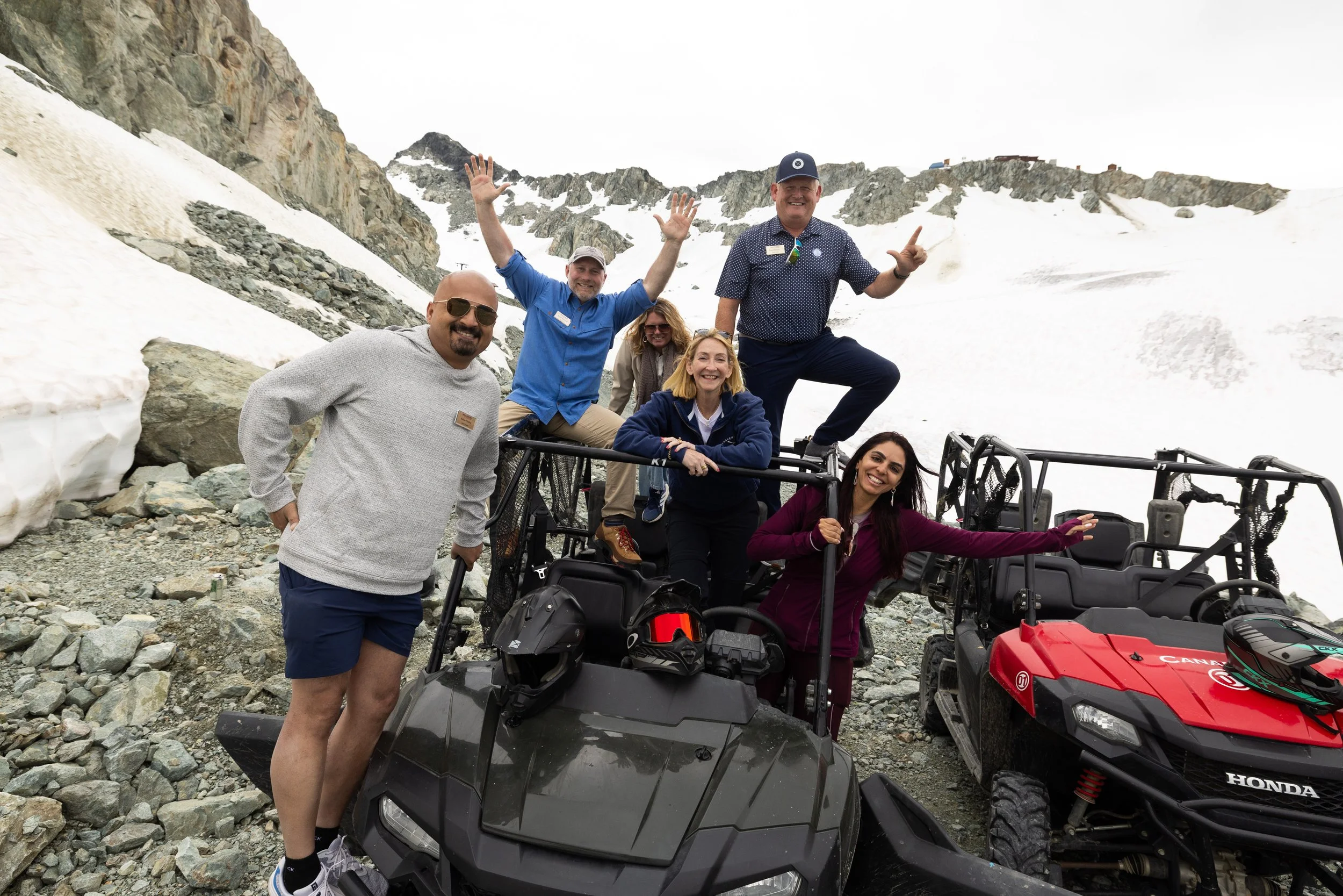 Group of six people on off-road vehicles in a mountainous, snowy landscape with rocks and snow patches.