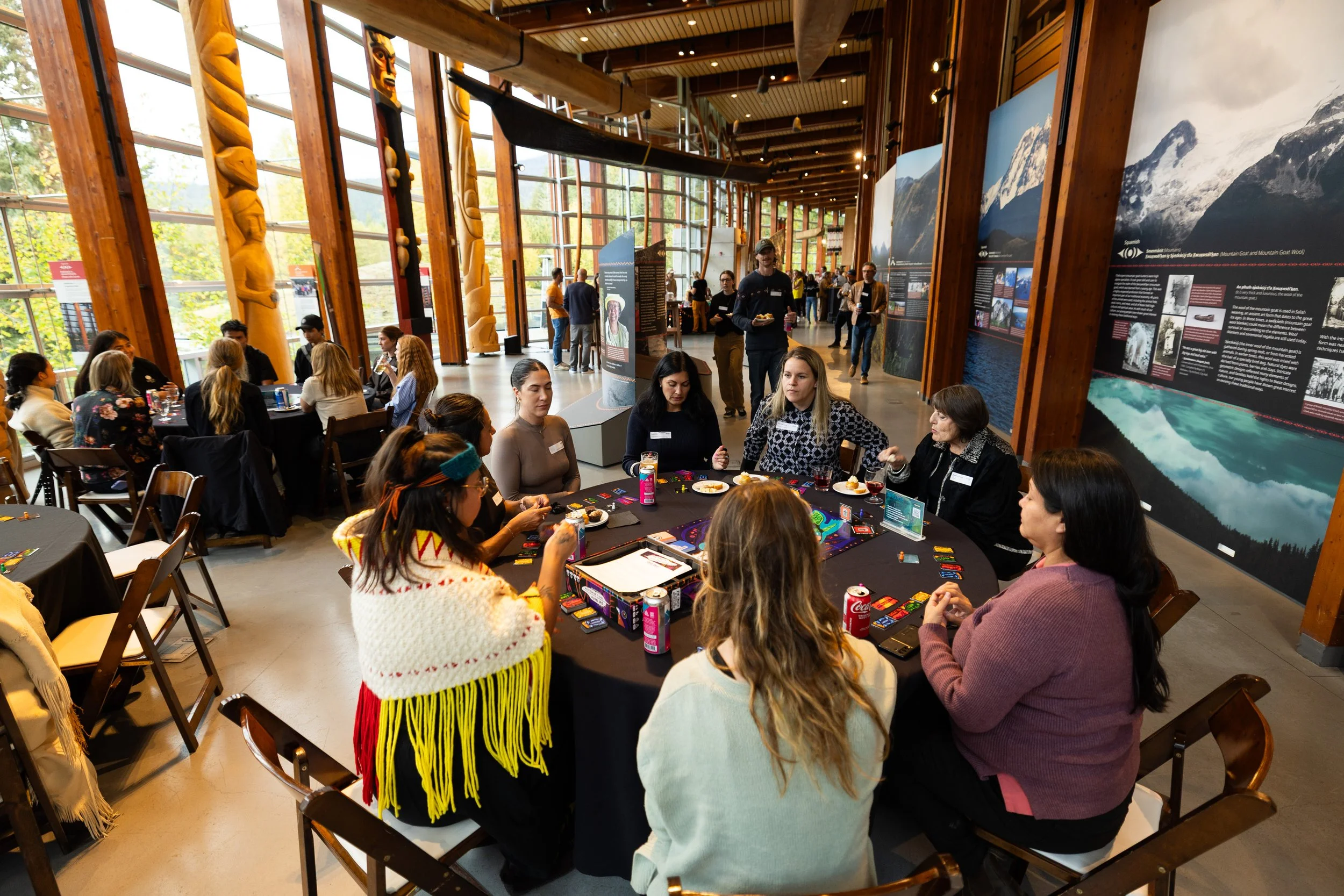 People sitting at round tables playing a card game in a spacious indoor setting with large windows and mountain-themed artwork on the walls.