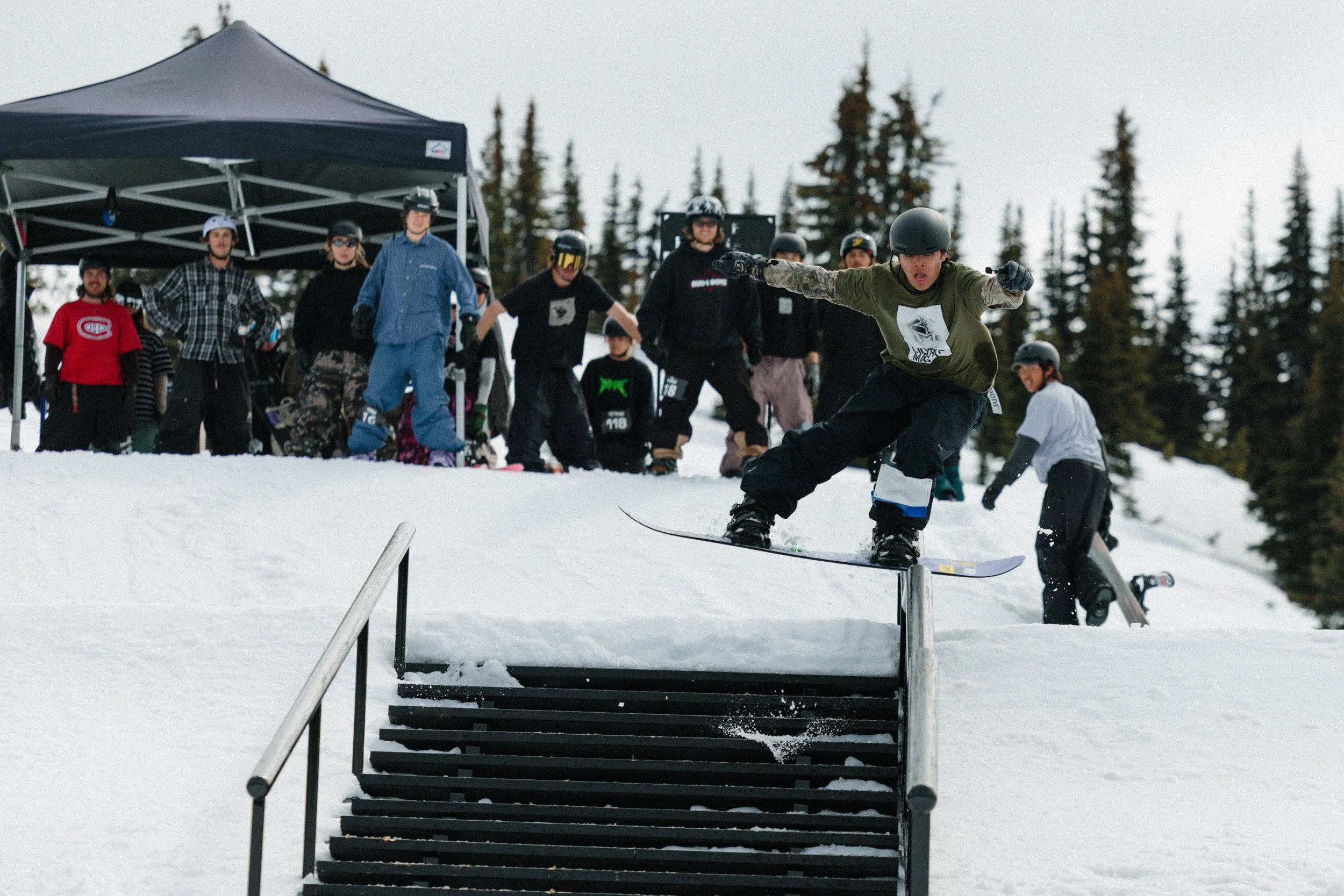 A group of people watch a snowboarder come down a rail at a snowy outdoor ski resort or park.