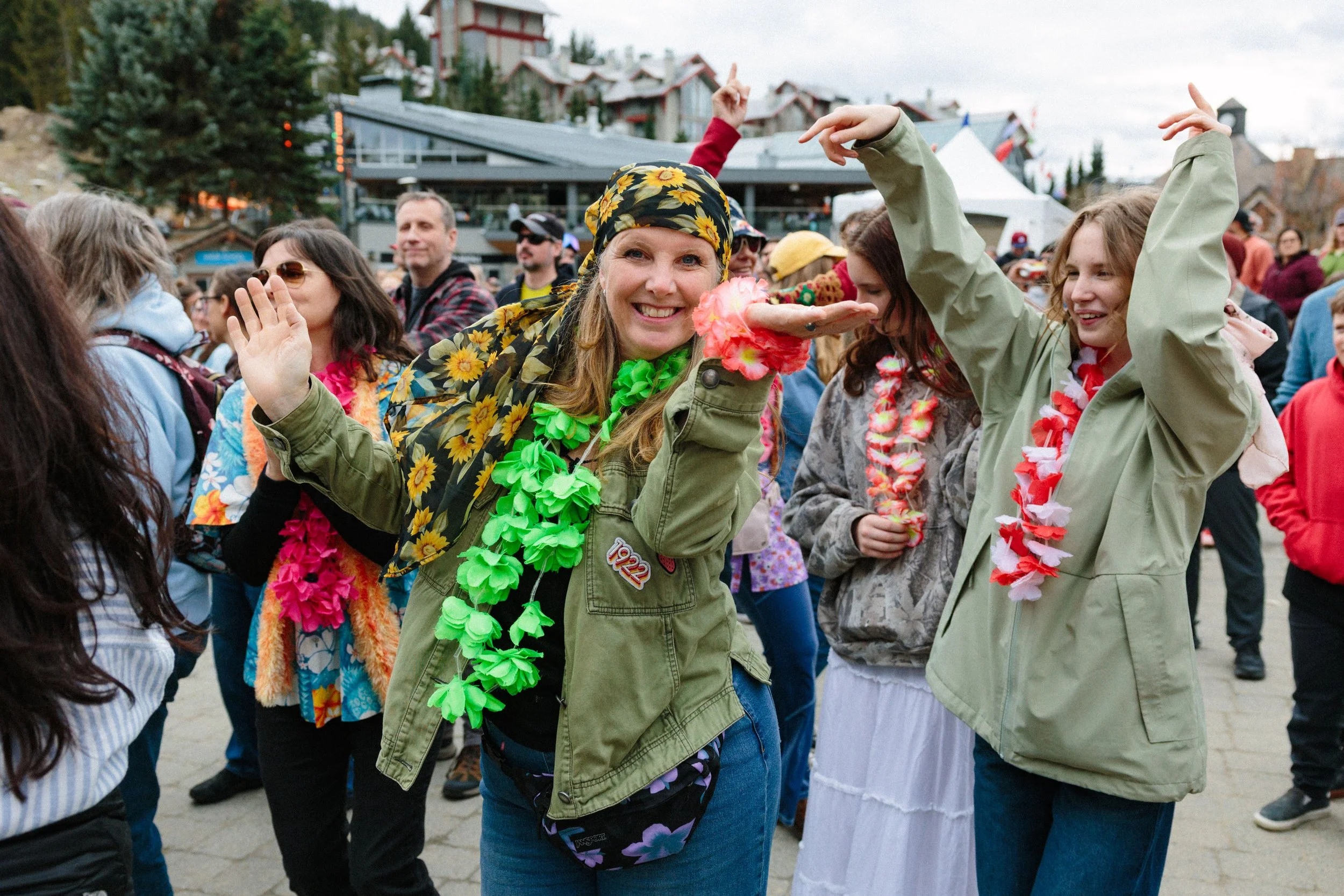 A group of people at an outdoor event, wearing colorful leis and Hawaiian-themed clothing. The woman in the center, smiling, has a bandana with sunflower print and a green lei, and nearby others are dancing or socializing.