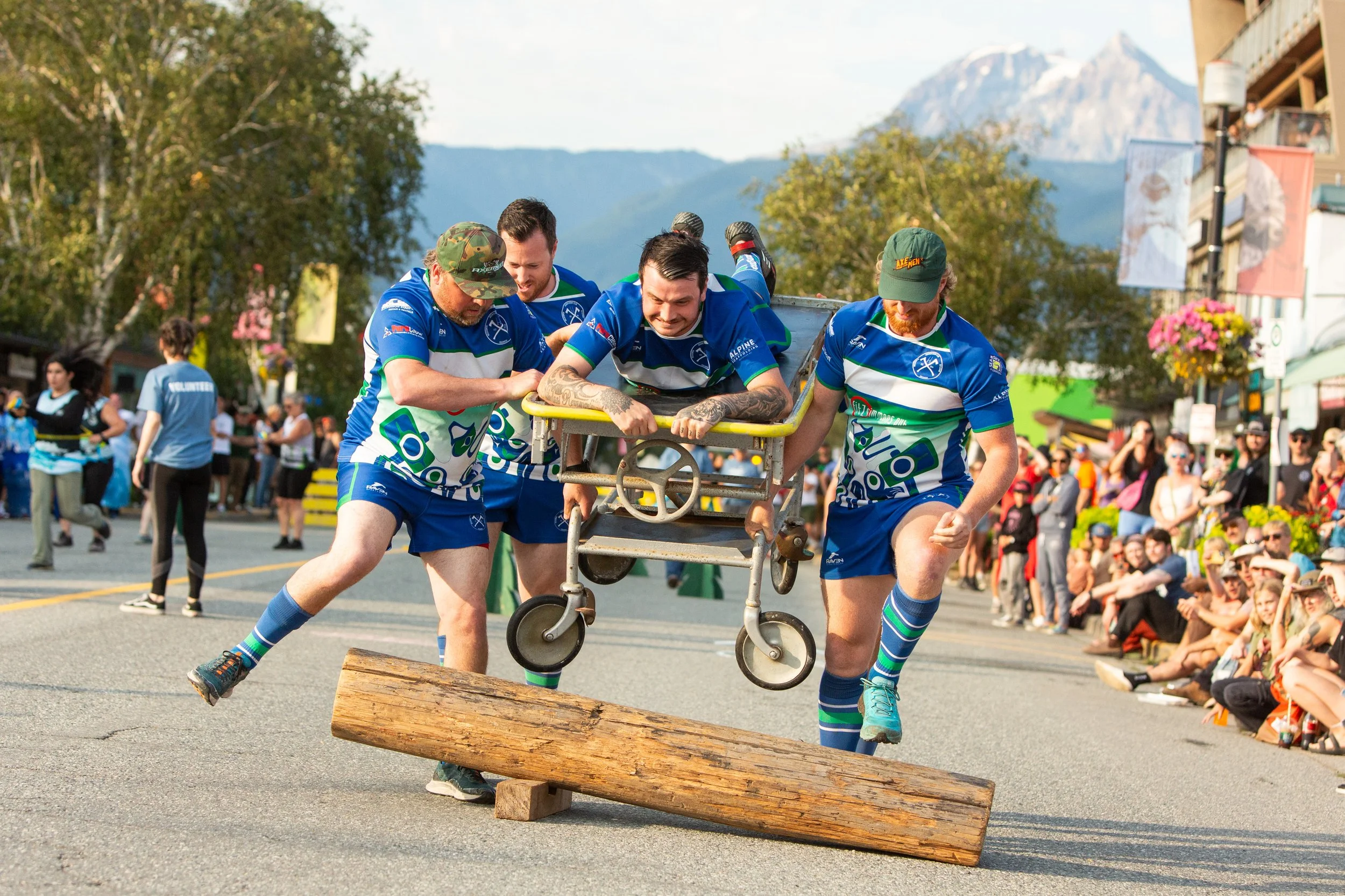 Four men in blue athletic uniforms are participating in a wheelbarrow race, carrying a person on a wheelbarrow over a logs obstacle, with a crowd and mountains in the background.