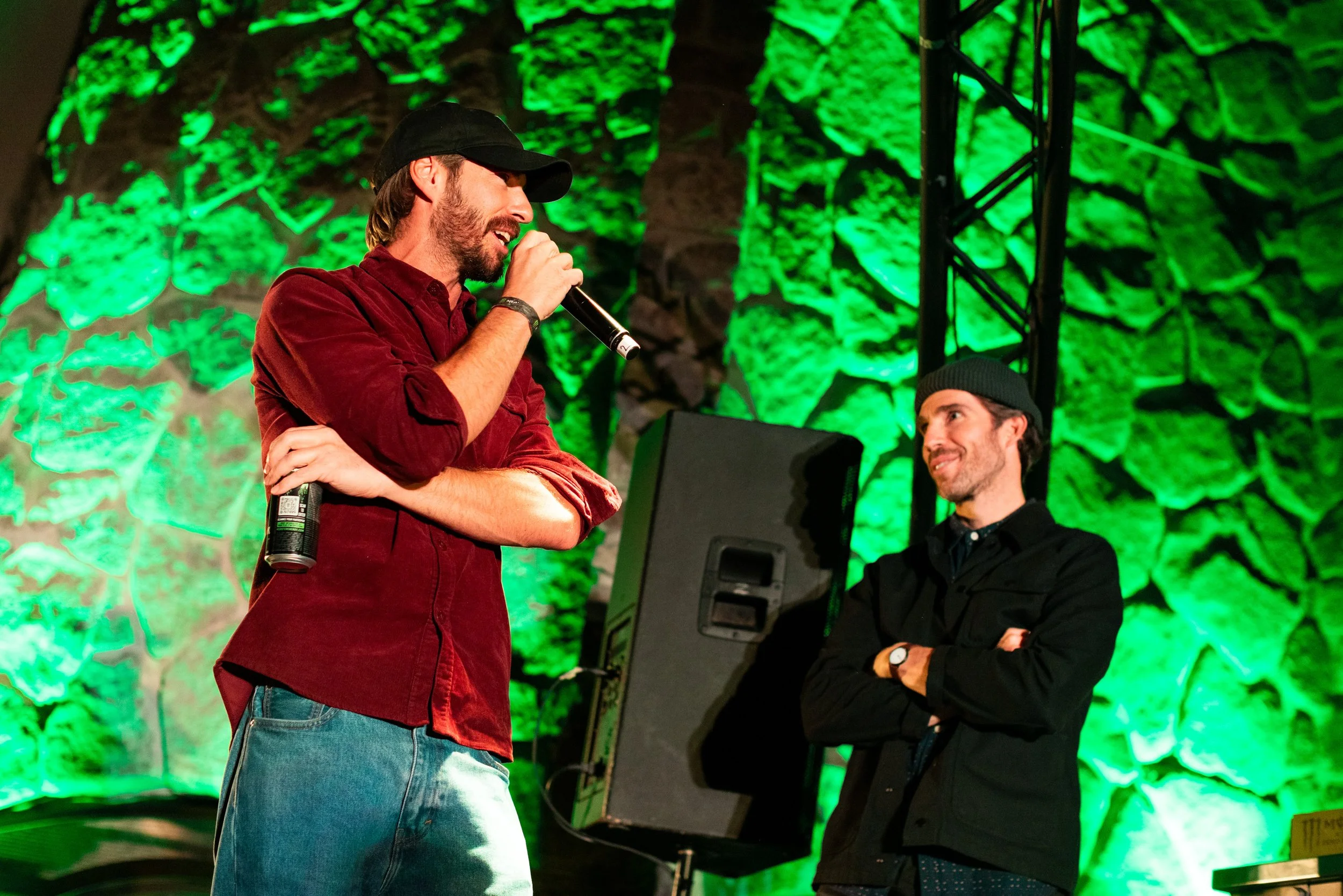 Two men are standing on a stage with a green lit stone wall background. One man, wearing a red shirt, blue jeans, and a black cap, is holding a microphone and speaking. The other man, wearing a black jacket and a black beanie, is smiling with arms cr