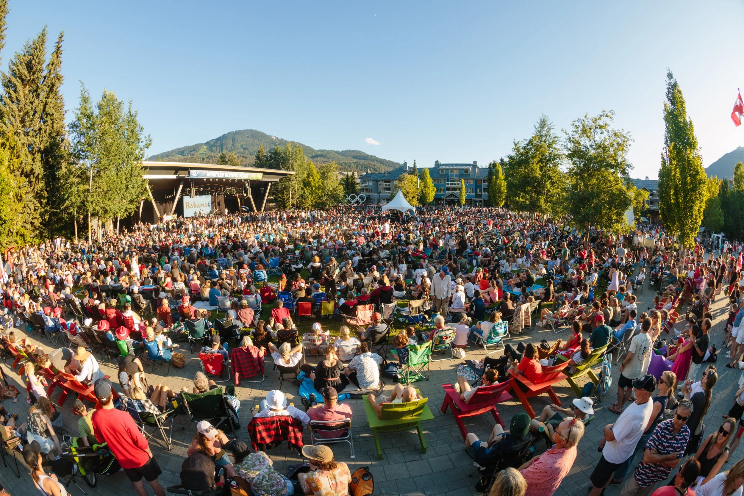 Large outdoor concert with a crowded audience seated on chairs and standing, facing a stage with a sign reading "Bahamas," in a park-like area with trees, mountains in the background, and a clear blue sky.