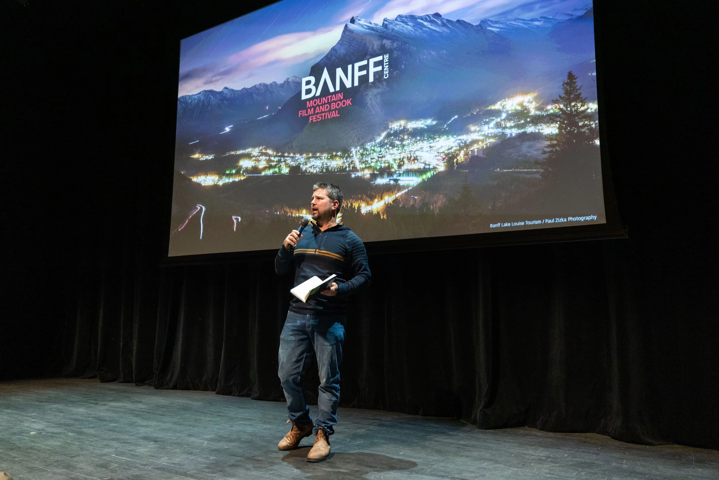 A man on stage holding a microphone and a notebook, presenting during the Banff Mountain Film and Book Festival. Behind him, a large screen displays a scenic nighttime view of Banff, with mountains, a town, and the festival's logo.