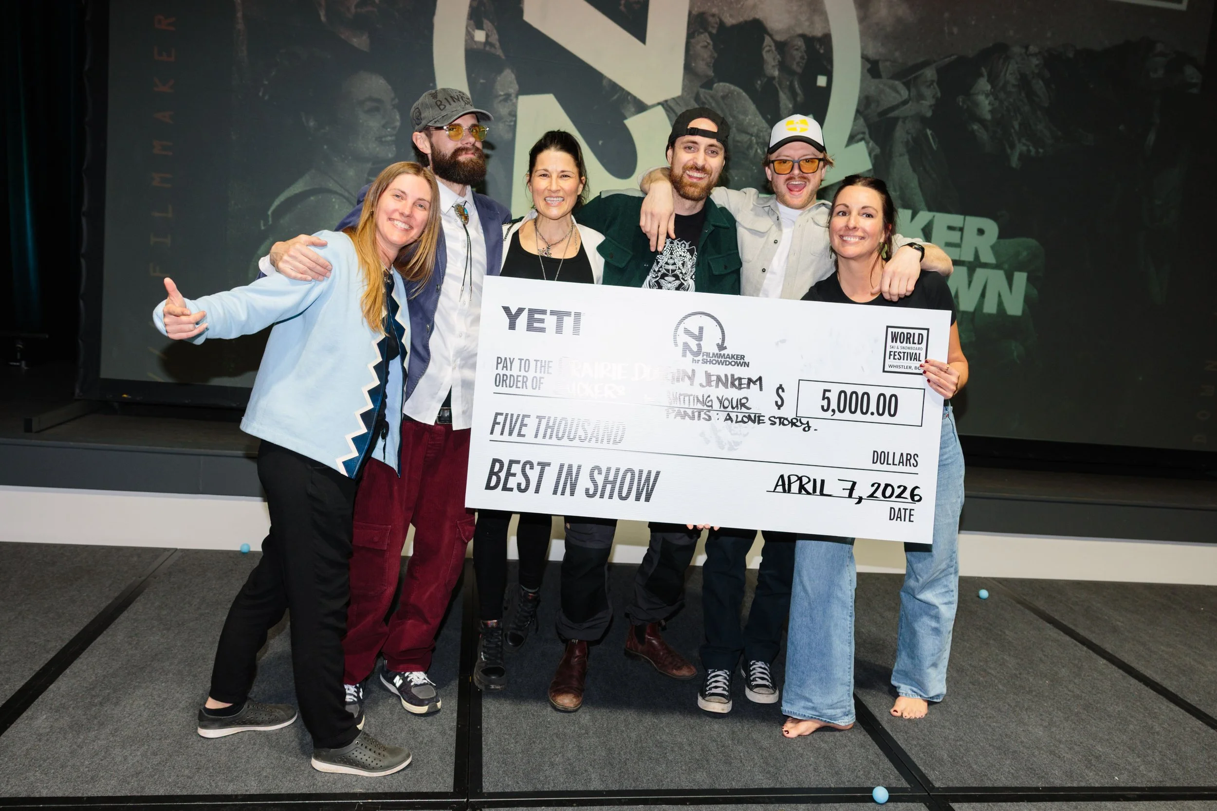 Group of six people at an awards event holding a large check with a background showing black and white images and a logo, celebrating their win.