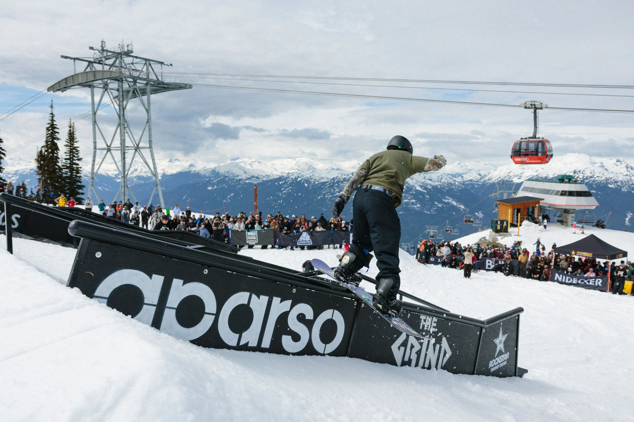 Skateboarder performing a trick on a rail at a snowy ski resort surrounded by crowds, mountains, and ski lifts.