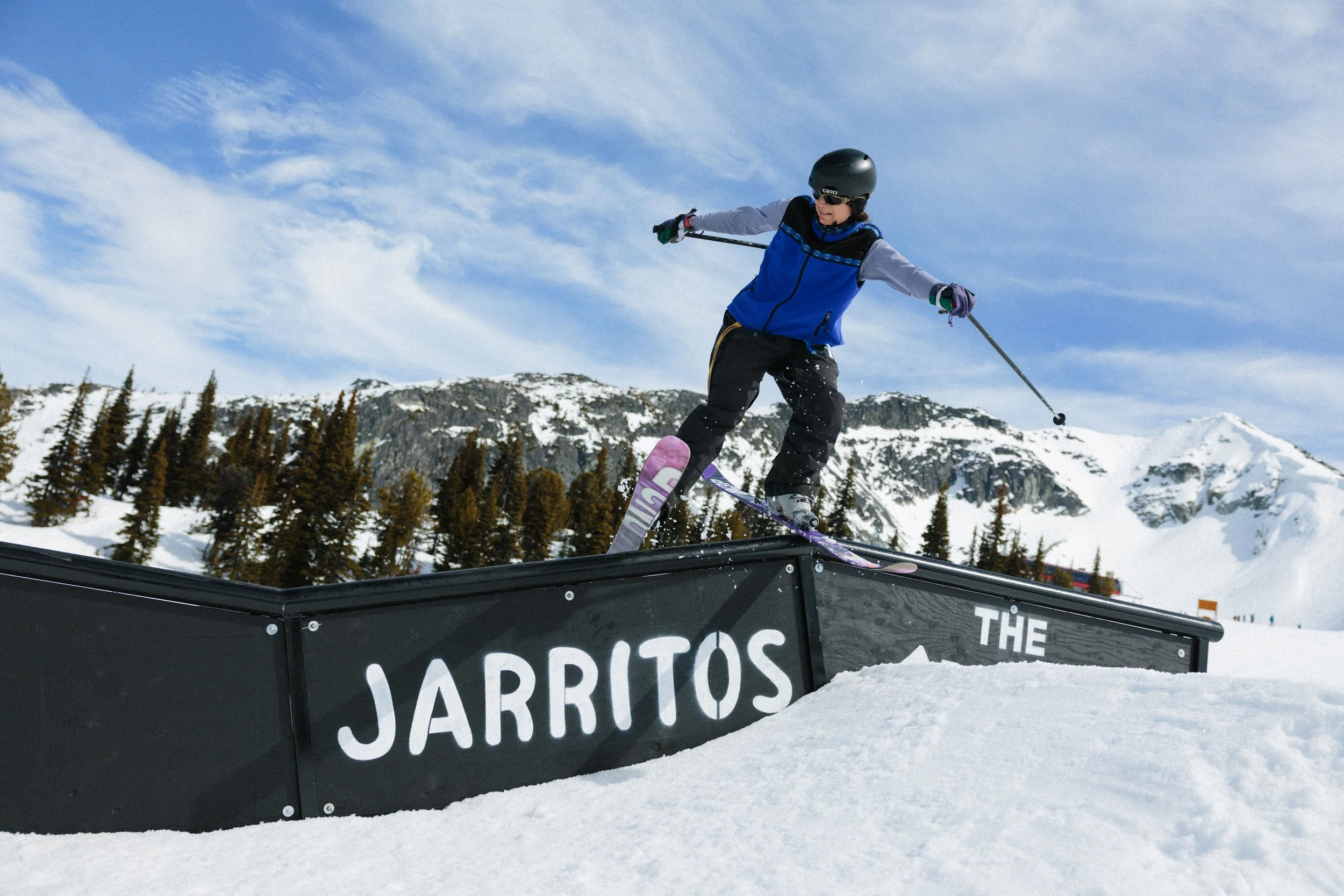 A person in winter gear, including a helmet and sunglasses, skiing down a snow-covered slope and jumping off a ramp in a mountain landscape with trees and rocky peaks under a blue sky.