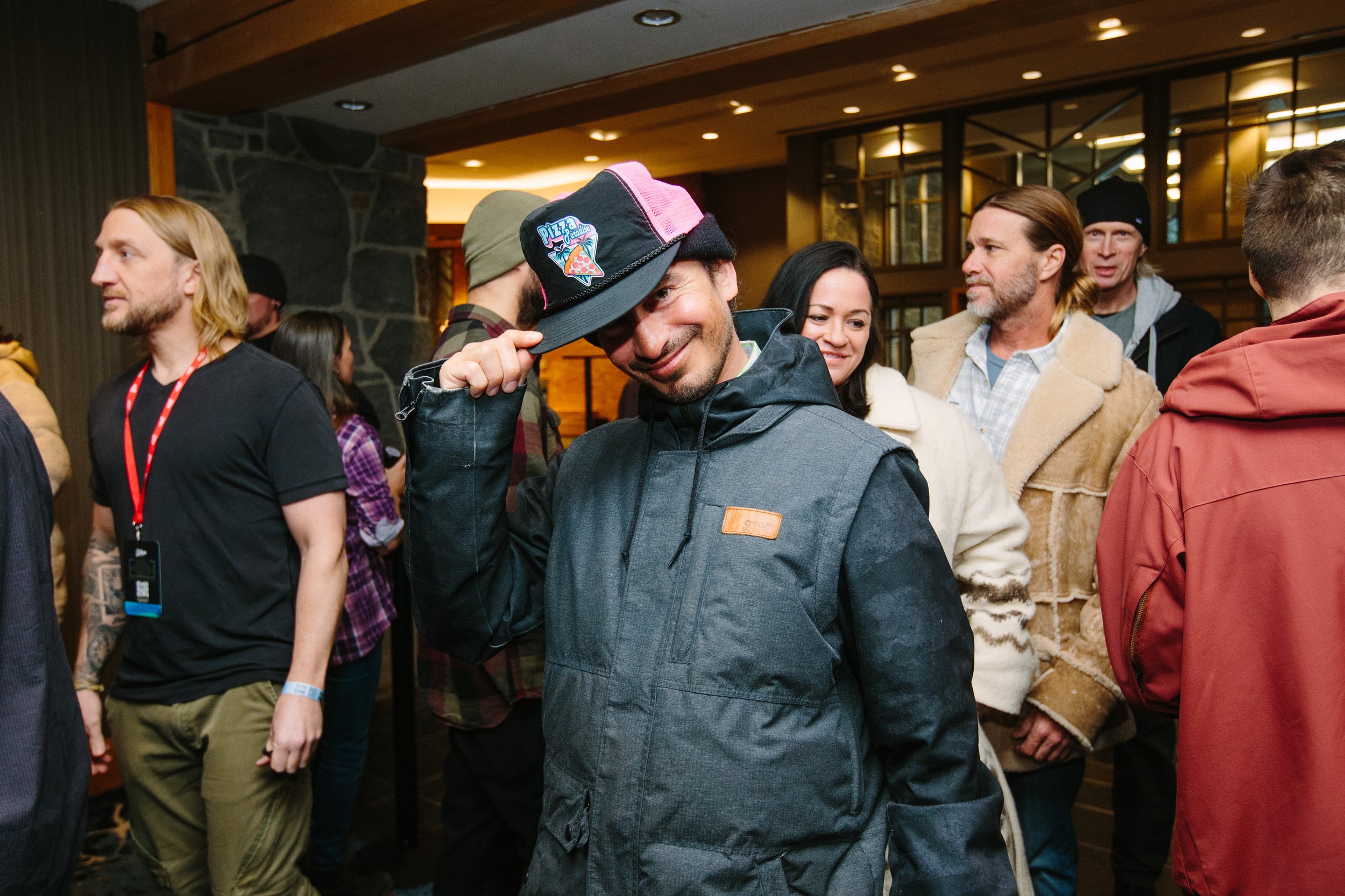 A man in a dark jacket and hat smiling and tipping his hat, surrounded by other people at an indoor social event.