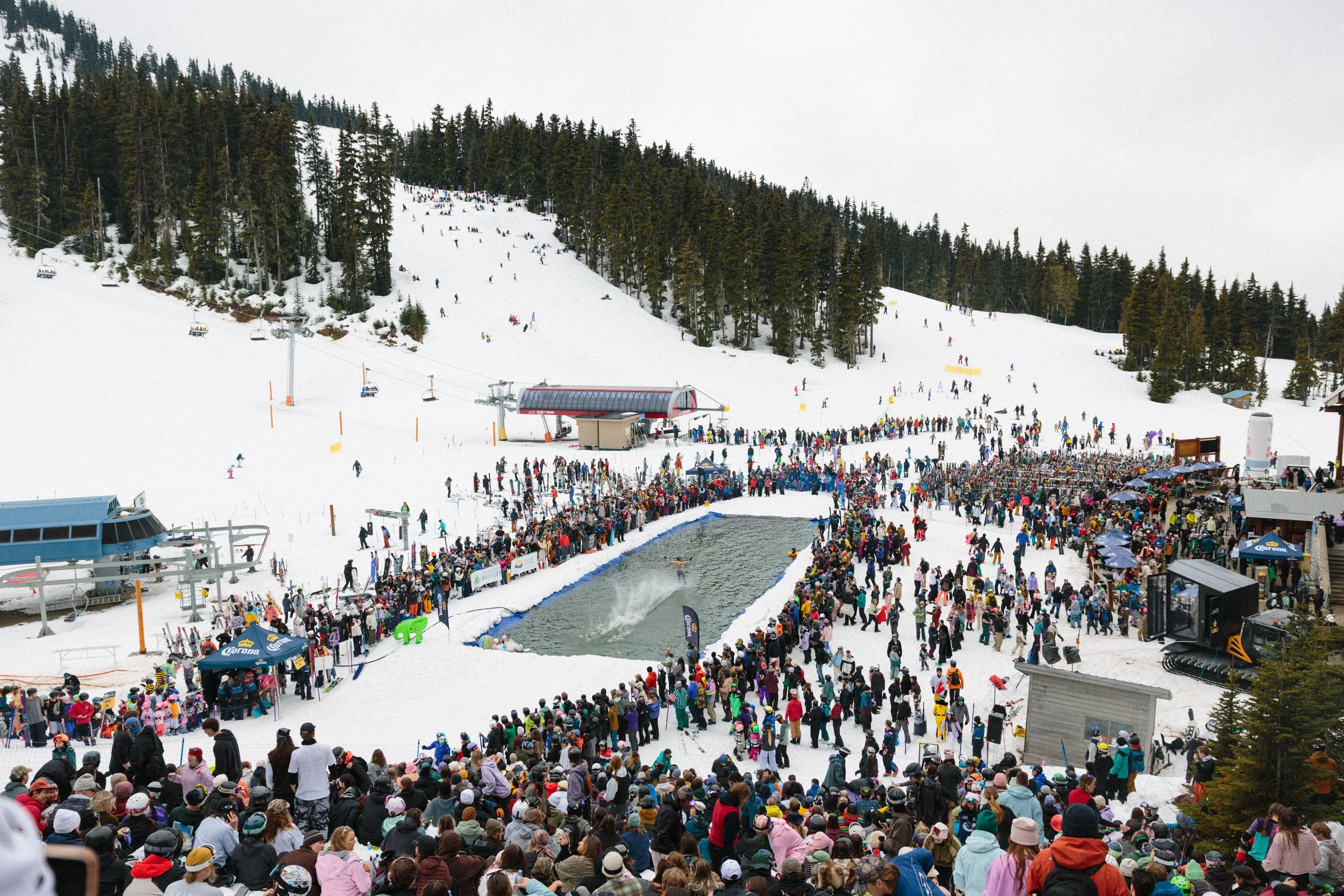 A crowded ski resort with many people waiting in line for a water-based ski jump in a snowy mountain landscape. There are ski lifts, a ski slope, and dense trees in the background.