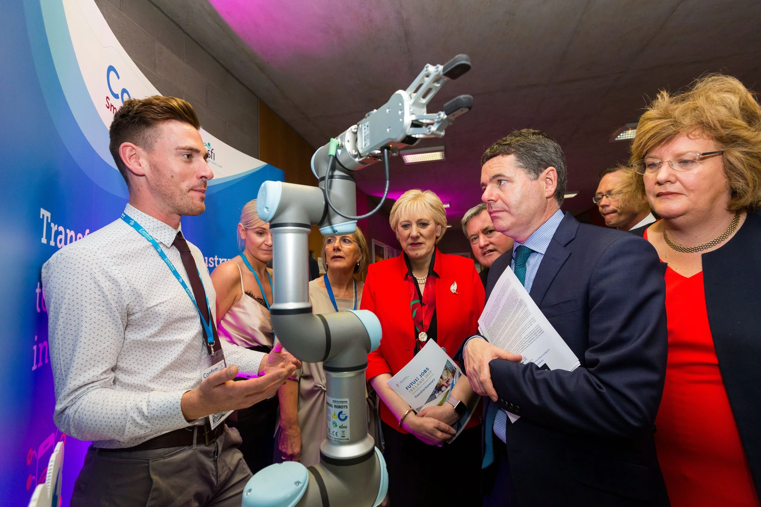 A group of professional-looking people, including men and women, gathered around a robotic arm at a conference or exhibition. One man is explaining something to the others, who appear attentive, with displays and banners in the background.