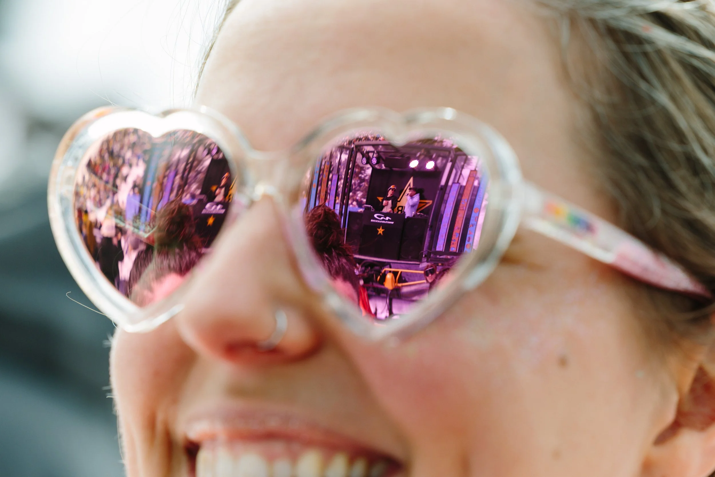 Close-up of a person's face wearing heart-shaped sunglasses, reflecting a stage with two performers, a DJ setup, and audience at an event.