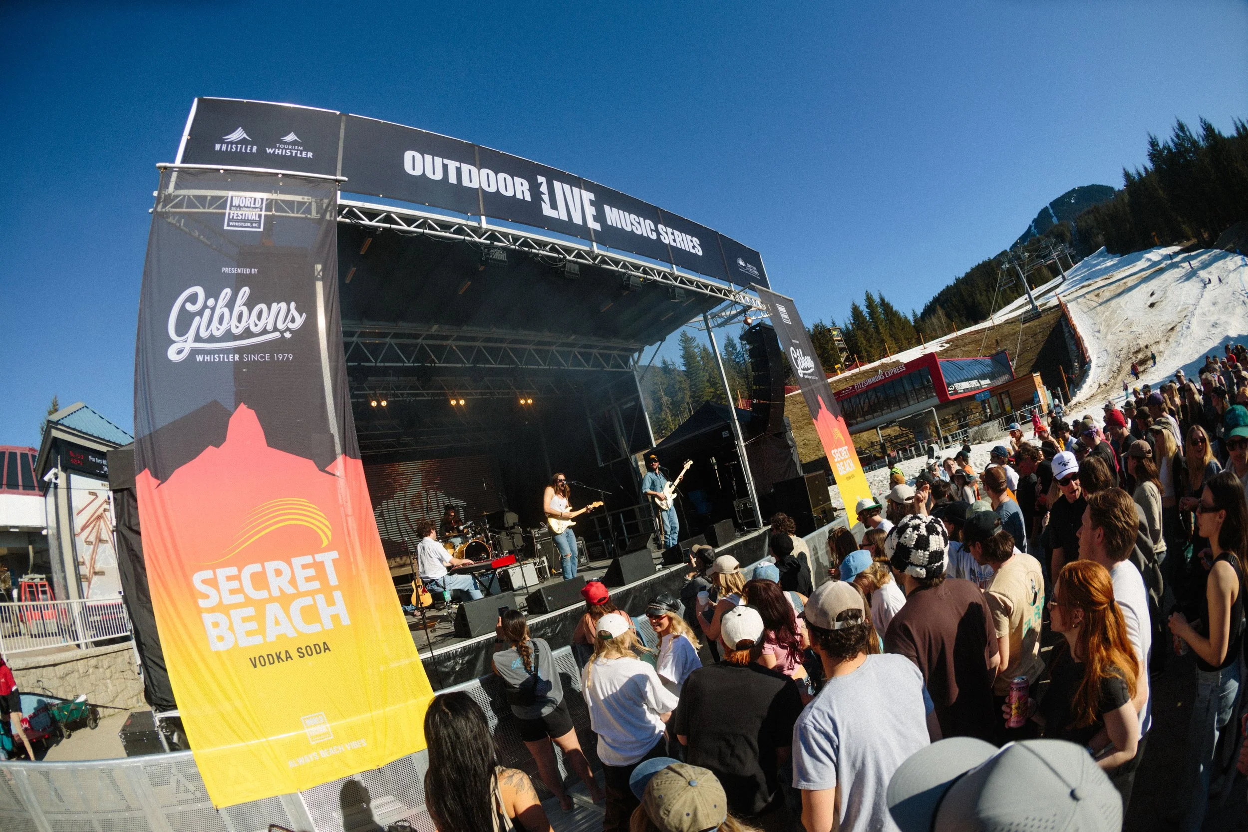 A large outdoor stage at the Whistler Outdoor Live Music Series during a festival, with a band performing. A crowd of people, some wearing hats and sunglasses, watch the concert. Colorful banners for 'Secret Beach' and sponsors are visible in the for