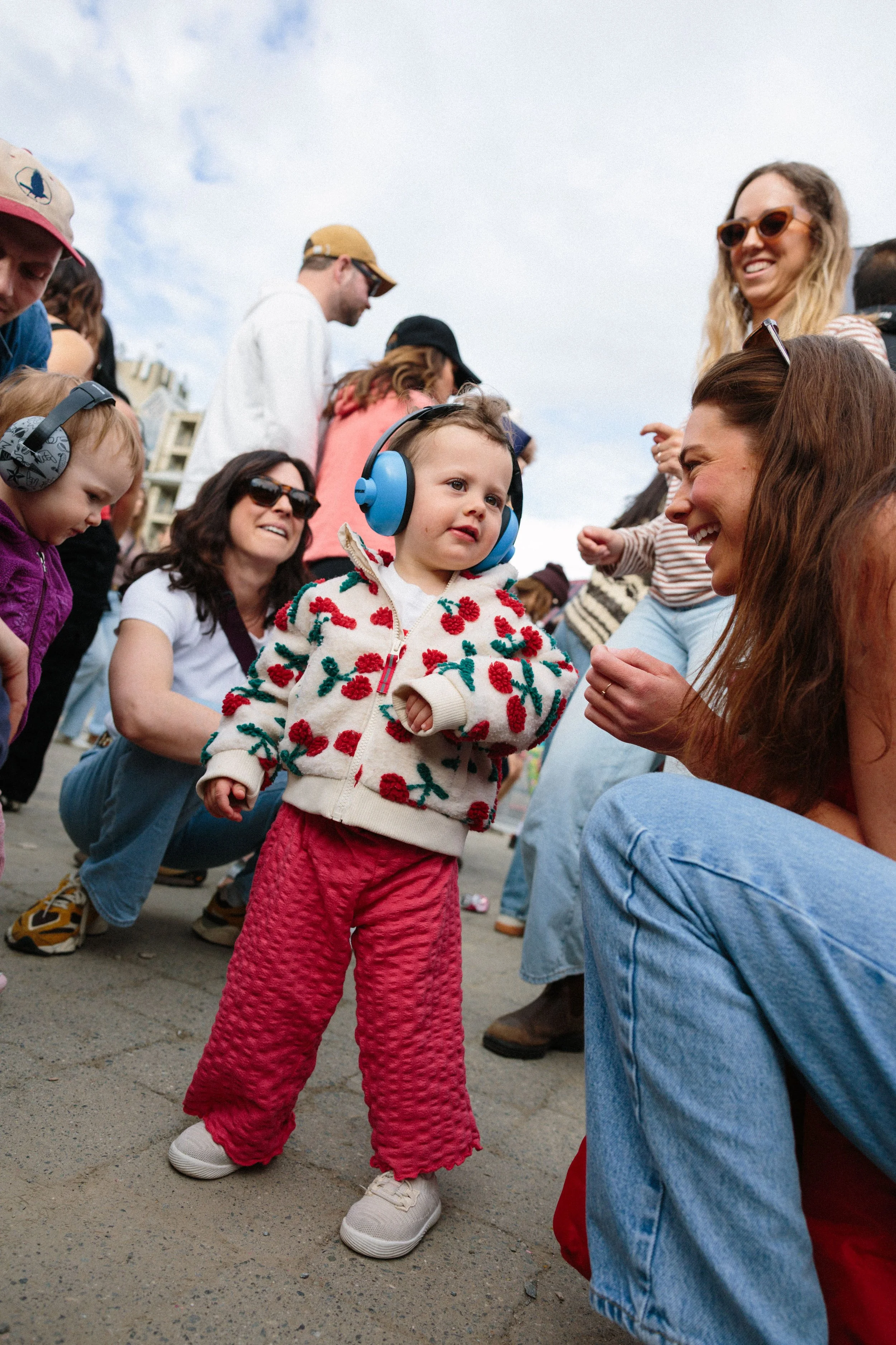A young girl with blonde hair is wearing large blue headphones, a cream cardigan with red and green floral embroidery, red quilted pants, and white sneakers. She is standing outdoors, smiling while surrounded by adults and children.
