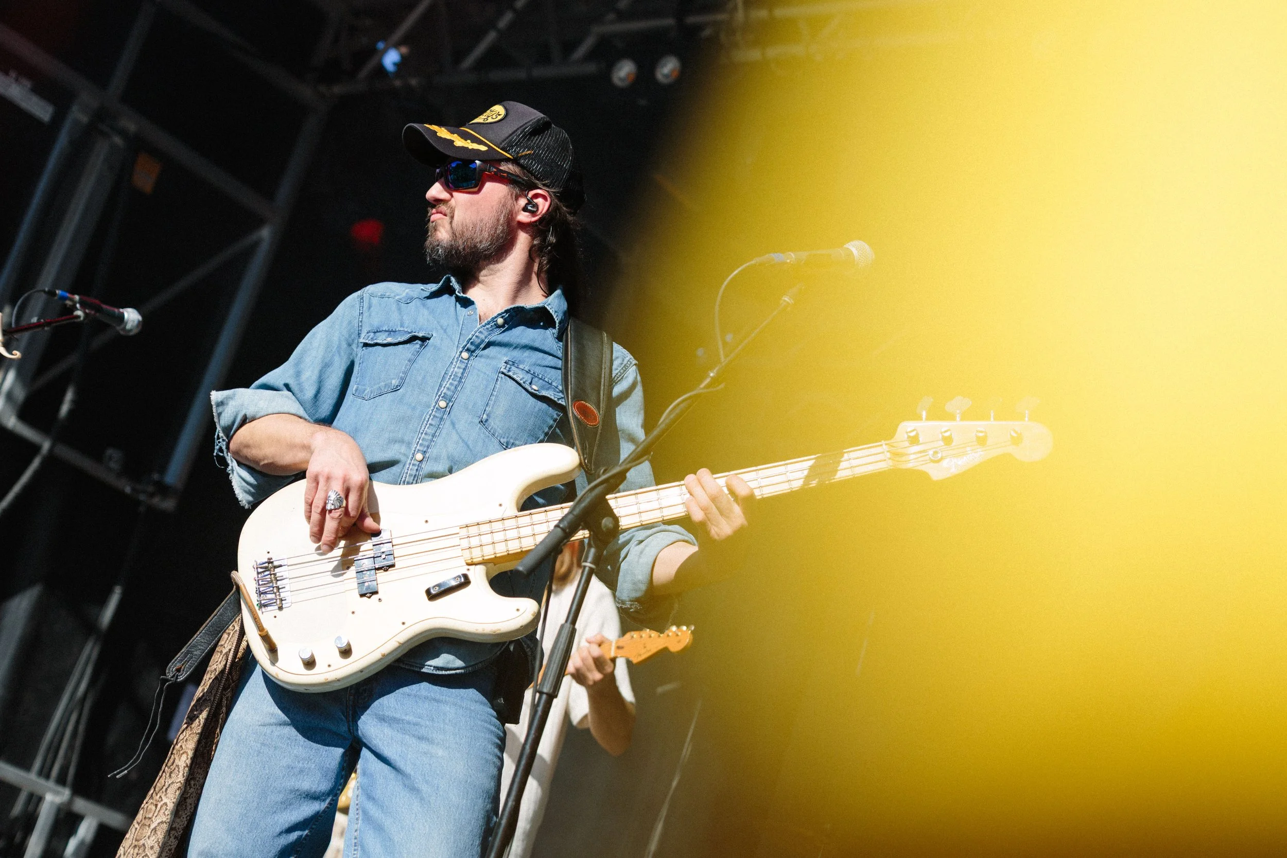 A man with long hair and beard wearing sunglasses, a black baseball cap, and a denim shirt, playing a white electric bass guitar during a live performance on stage with bright yellow lighting.