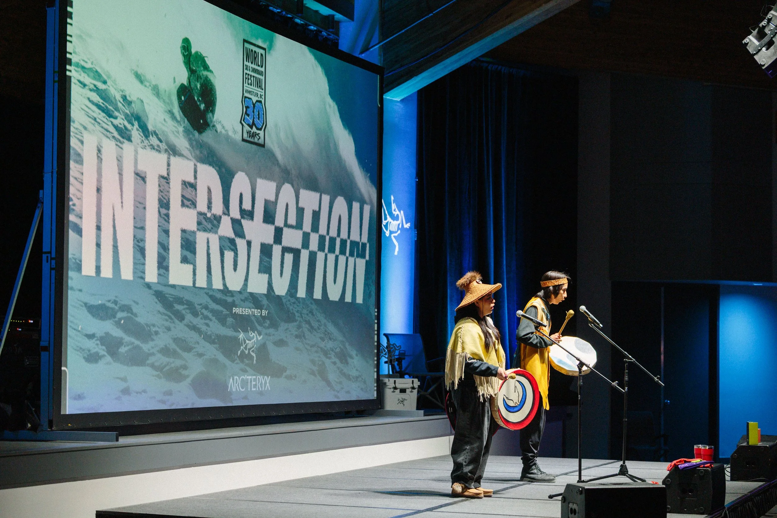 People dressed in indigenous clothing playing drums and holding shields on stage at a festival, with a large screen displaying the words "International" and a logo celebrating 30 years of the World Steal Snowboard Festival in Whistler, BC.