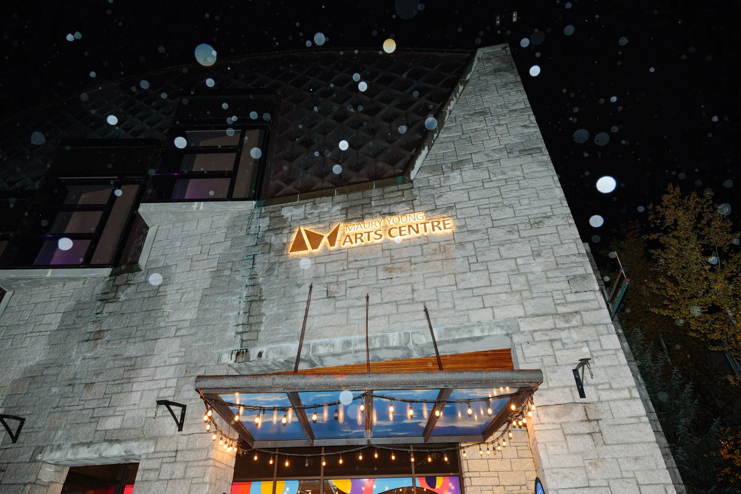 Nighttime exterior view of the Maury Young Arts Centre with a stone facade, illuminated sign, and a window with string lights hanging above the entrance.