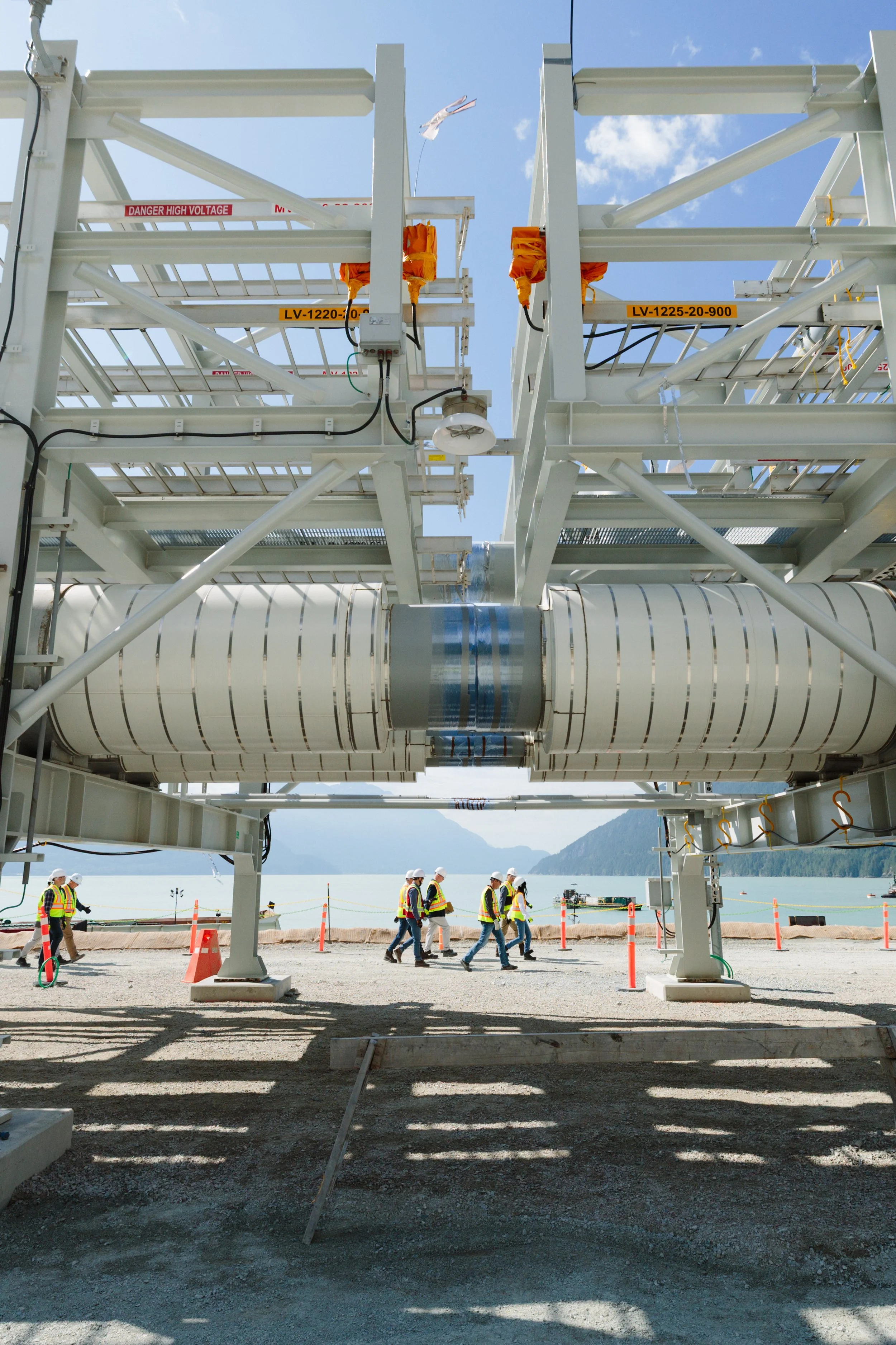 Construction workers walking on a gravel surface beneath large industrial machinery at a waterfront construction site with mountains in the background.