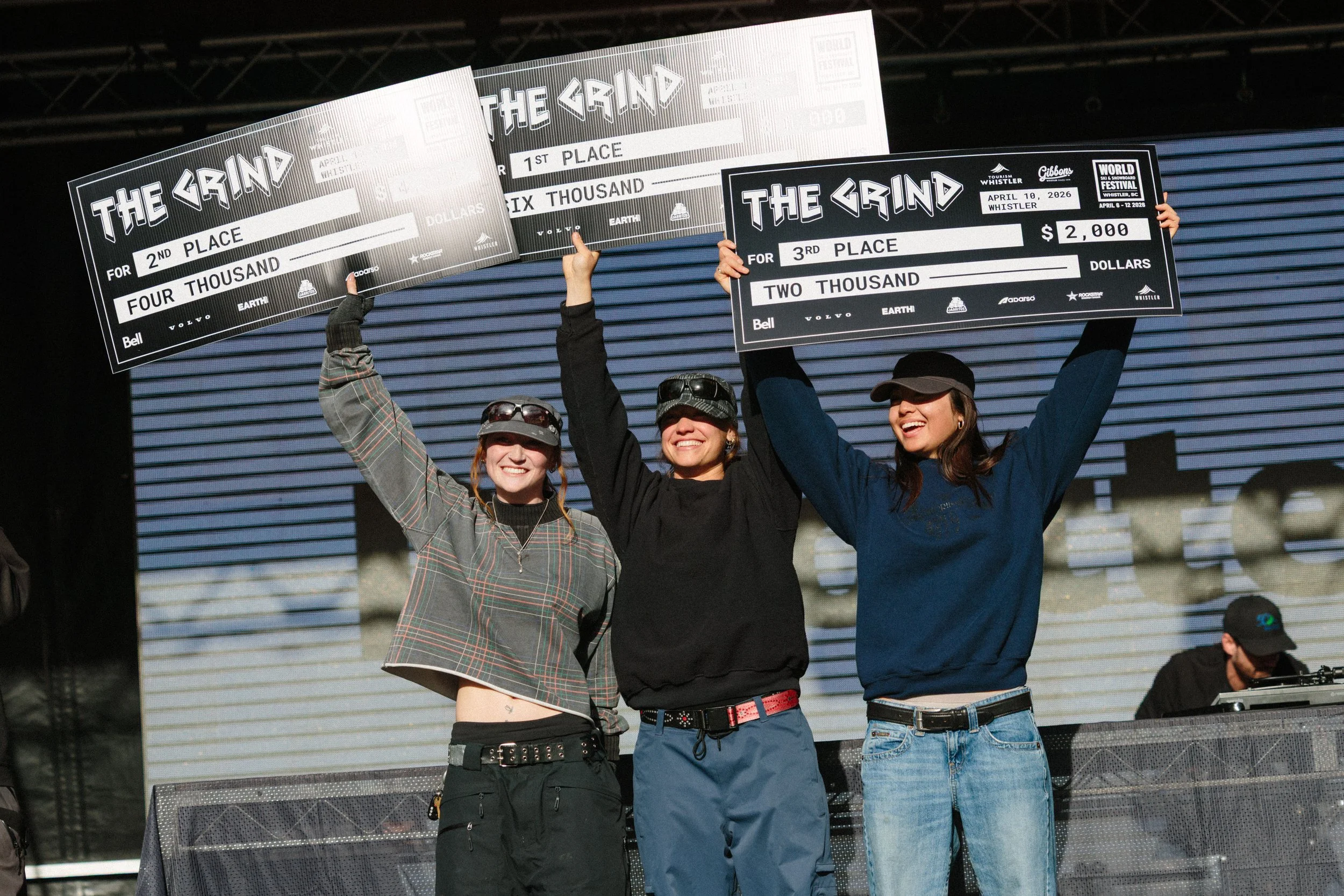Three women standing on a stage, holding large check awards for first, second, and third place at a competition called 'The Grind.' The checks display the location as Whistler, BC, and the date as April 10, 2026. Each woman is smiling and dressed cas