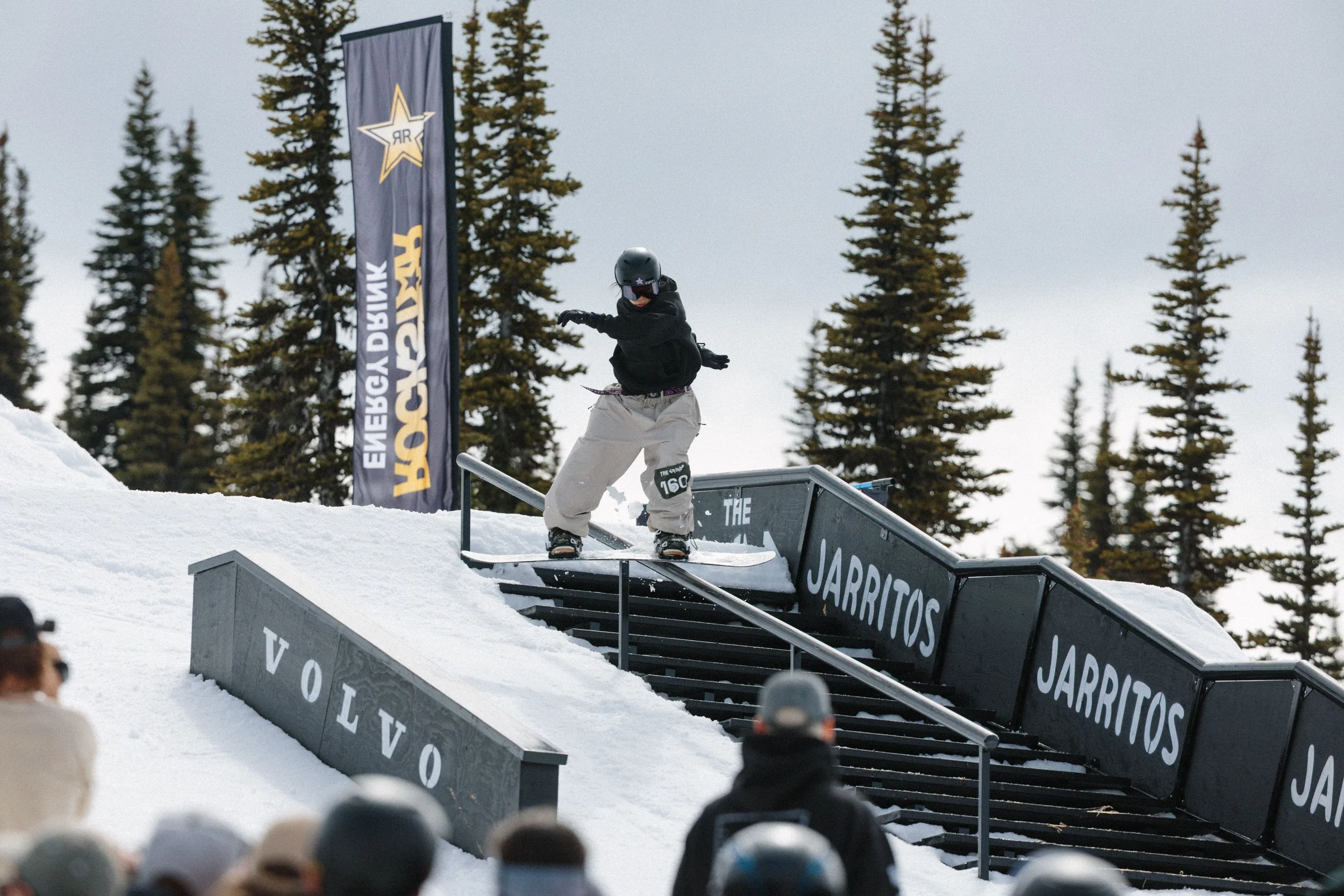 Skateboarder in black helmet and black jacket performing a rail slide on a rail at a snowboarding park, with spectators in the foreground and tall pine trees in the background.