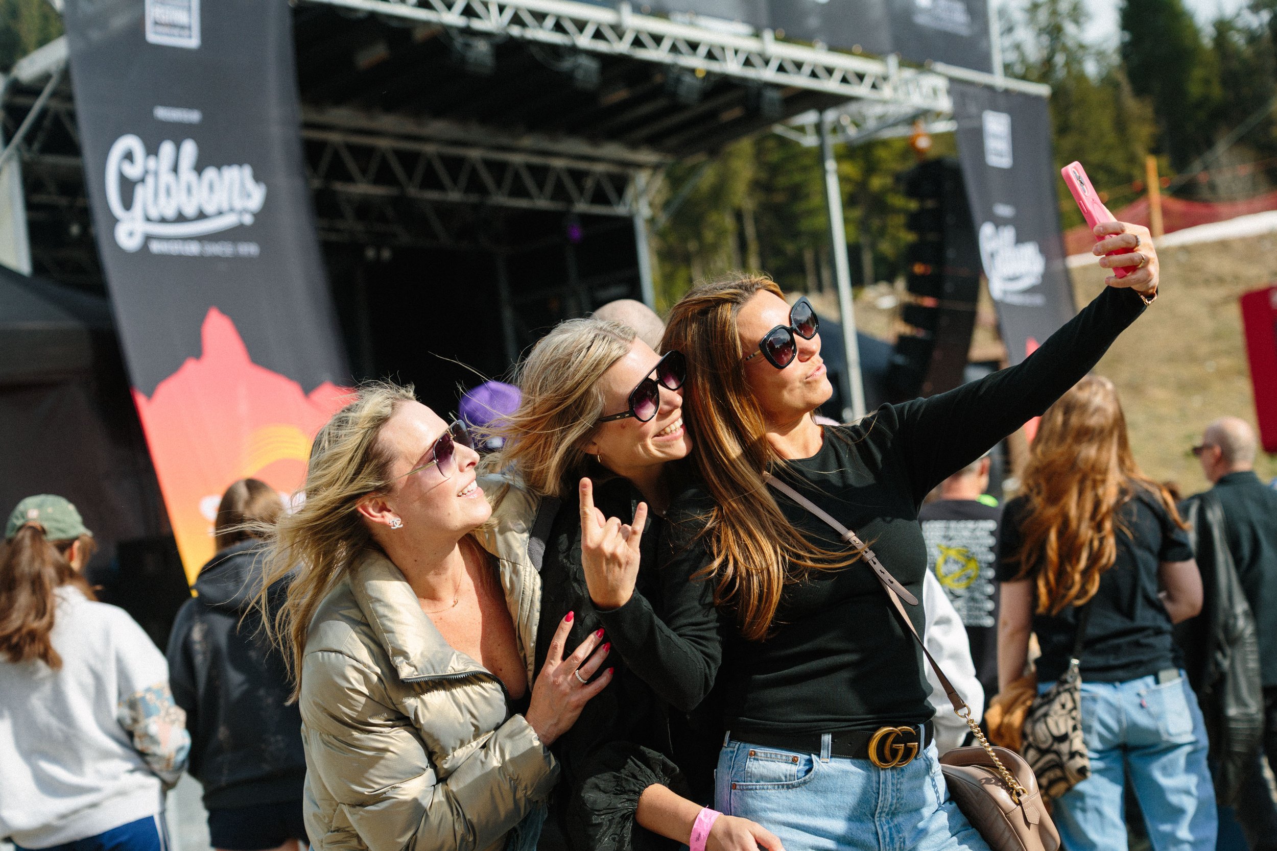 Three women wearing sunglasses taking a selfie at an outdoor event with a crowd in the background, and a stage with a black backdrop that has a logo and text.