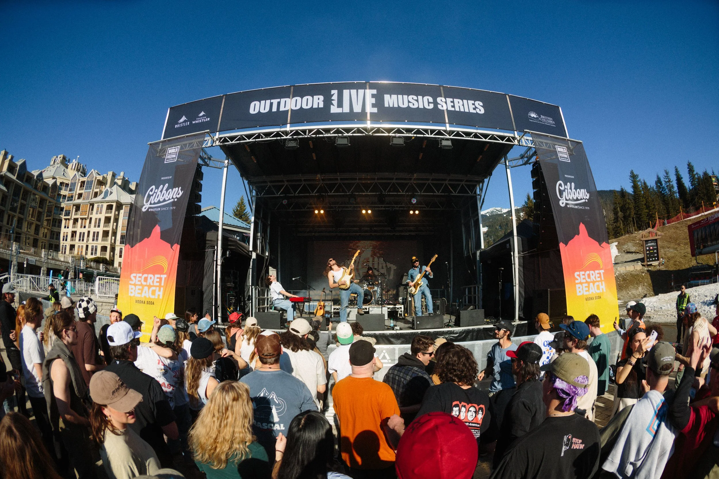 An outdoor stage at a music festival with a band performing, surrounded by a crowd of people wearing hats and casual clothing, with mountains and buildings in the background on a clear blue day.