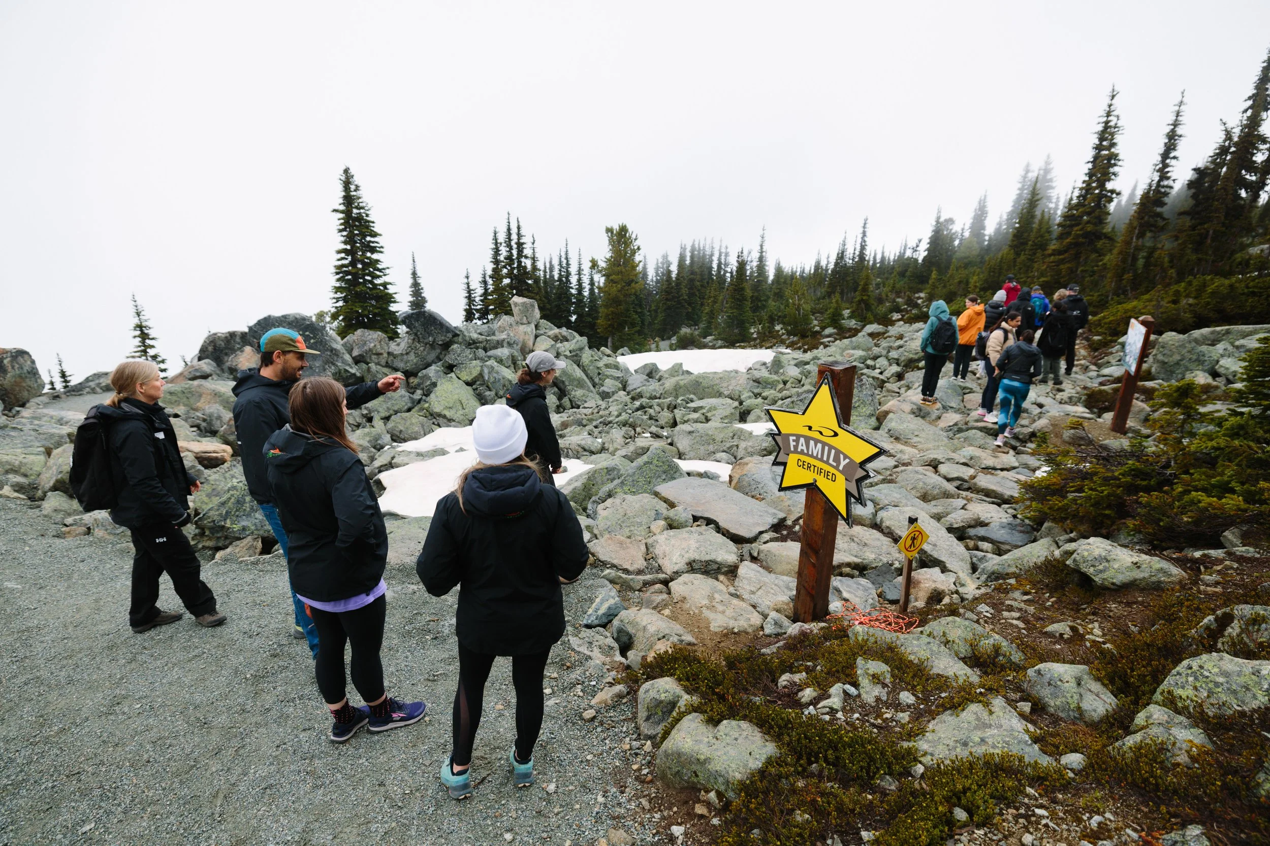 Group of people on a rocky mountain trail, some looking and others walking. There are warning signs, including one indicating 'Family Certified'; the sky is overcast and snow patches are visible.