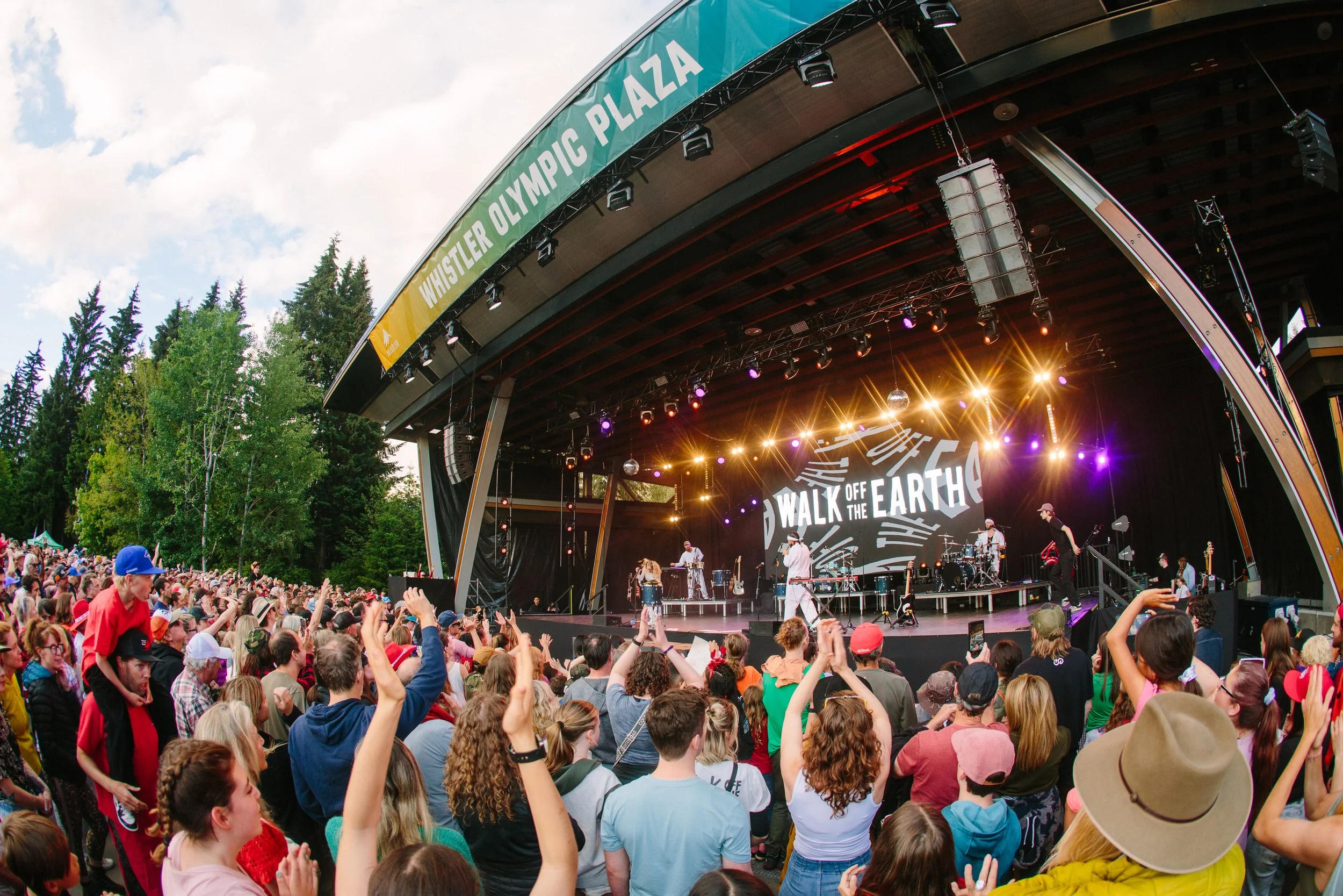Outdoor music concert with a large crowd gathered in front of a stage with performers and a banner reading "Whistler Olympic Plaza" and "Walk Off The Earth".