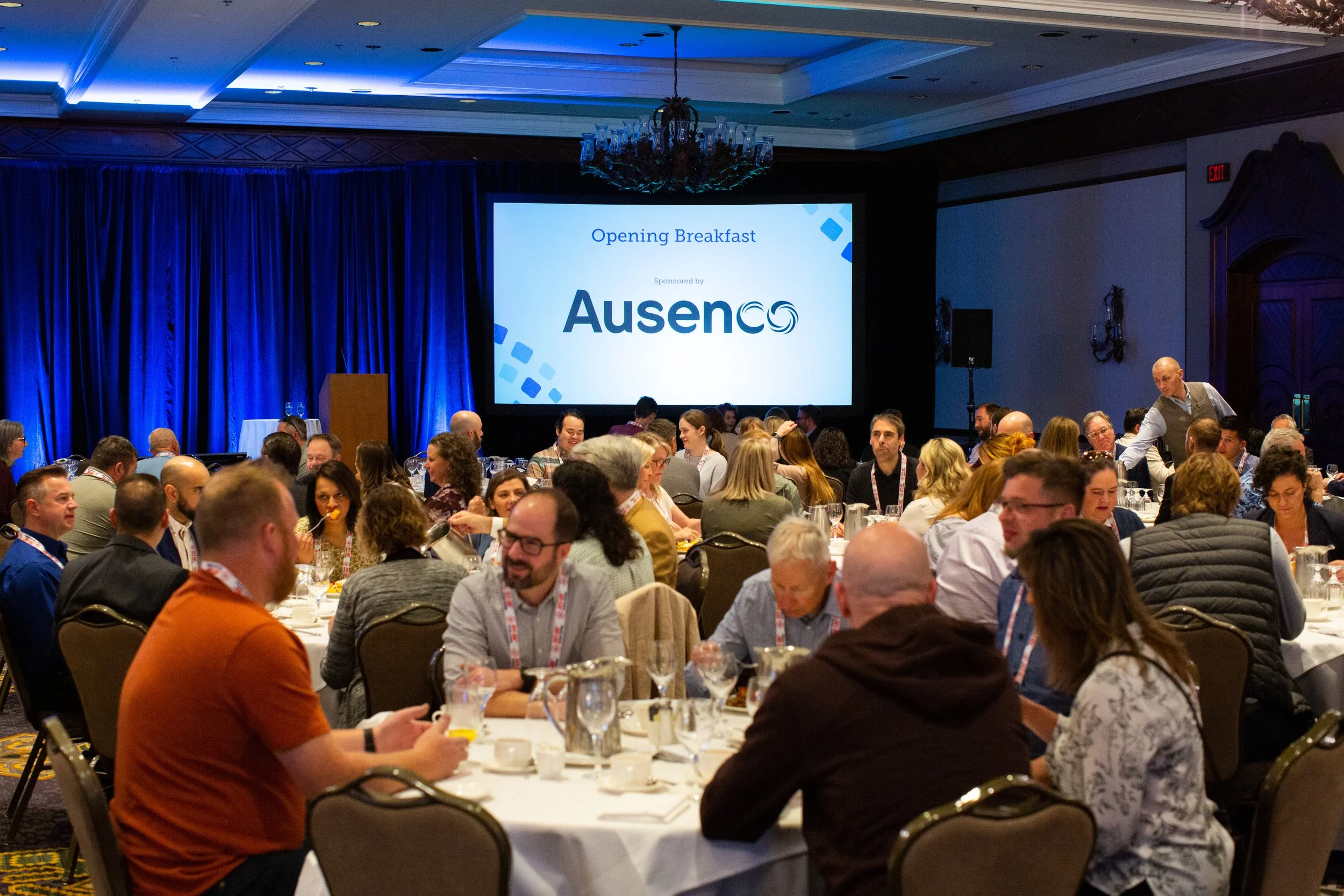 Conference attendees seated at tables during breakfast event sponsored by Ausenco, with a screen displaying "Opening Breakfast."