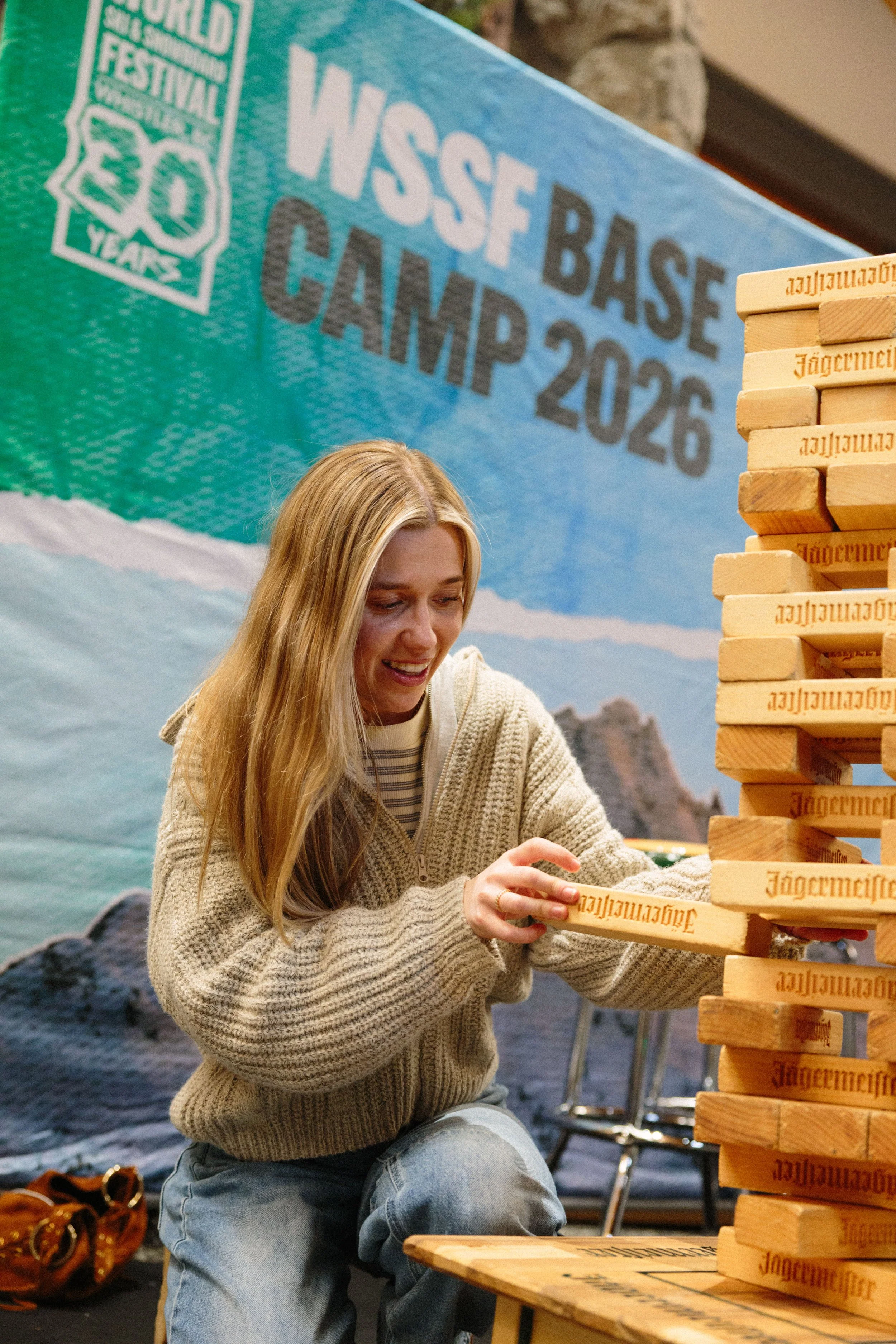 A young woman in a beige sweater and blue jeans playing Jenga at an indoor event with a backdrop featuring a mountain and a green banner that says 'WSSF Base Camp 2026'. She is smiling and focused on removing a piece from the Jenga tower.