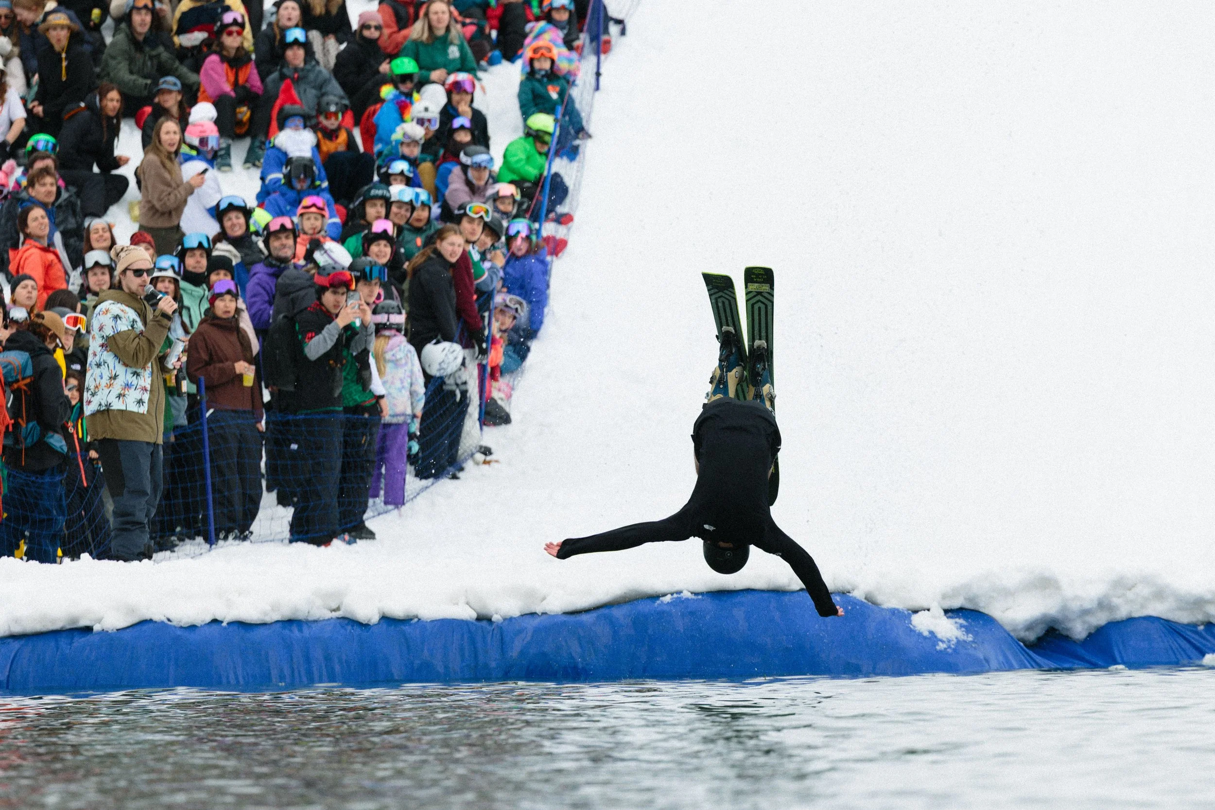 A person performing a handstand on snow with skis on their feet at a ski event, spectators bundled in winter clothes watching from behind a fence.